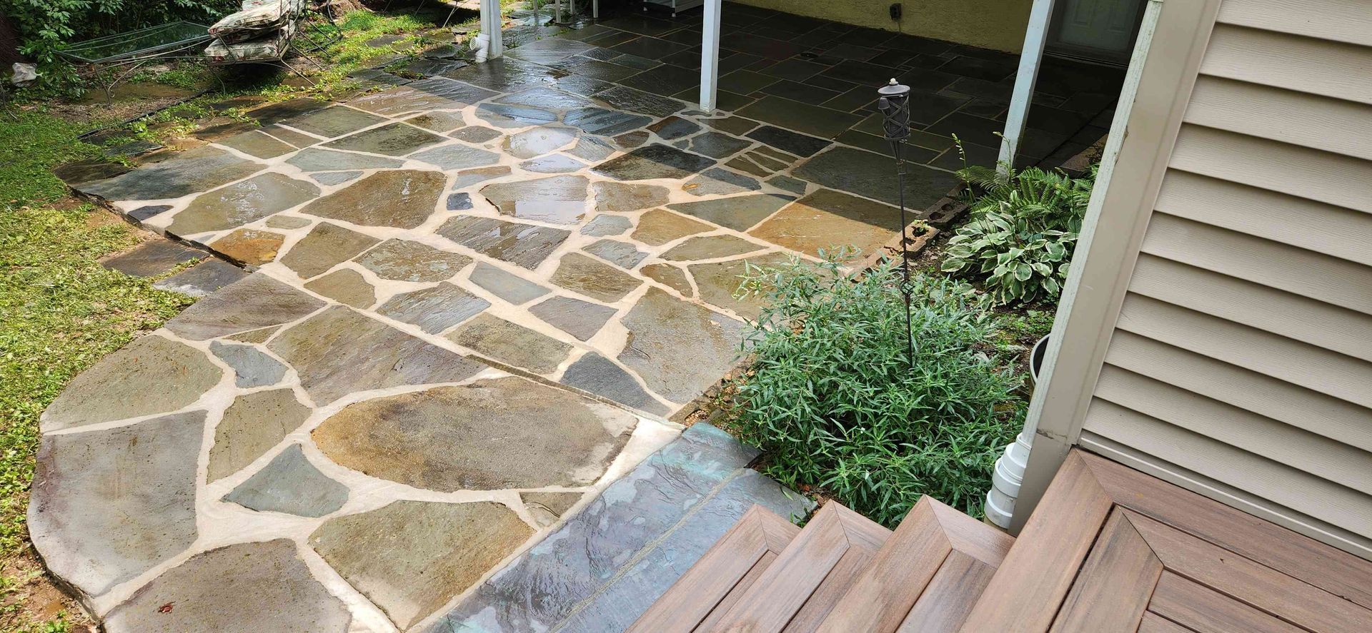 A flagstone patio featuring irregular tan and grey stones with light grout, adjacent to wooden steps and green bushes.