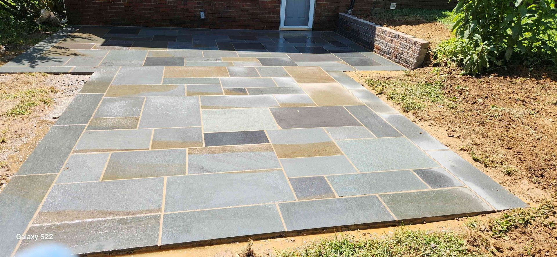 A gray rectangular stone patio with a natural pattern, bordered by unfinished soil and a brick wall.