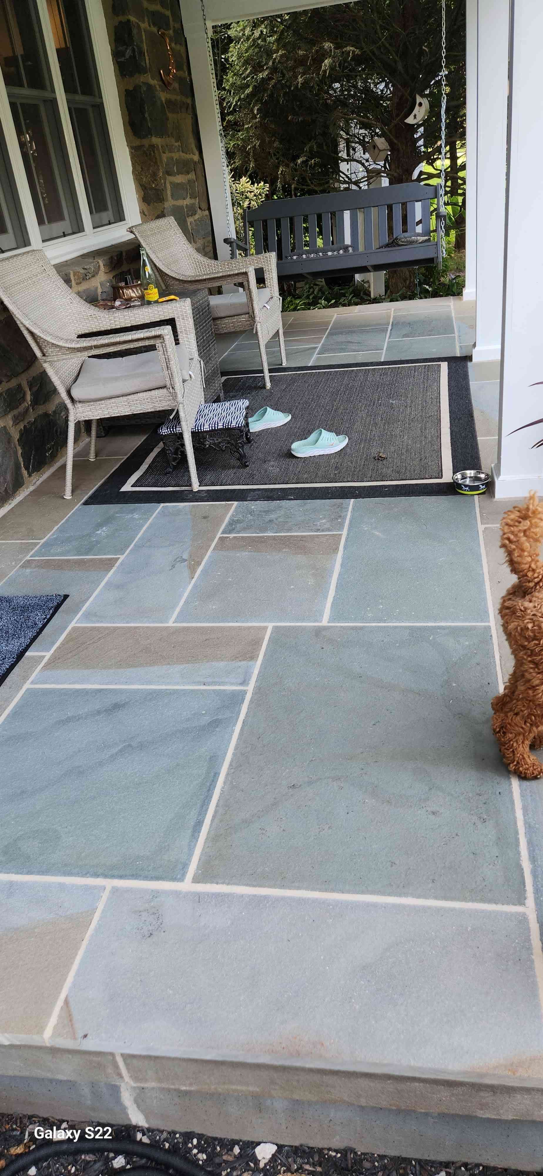 A stone-paved front porch with wicker chairs, a rug, and a small, curly-haired puppy standing to the right.