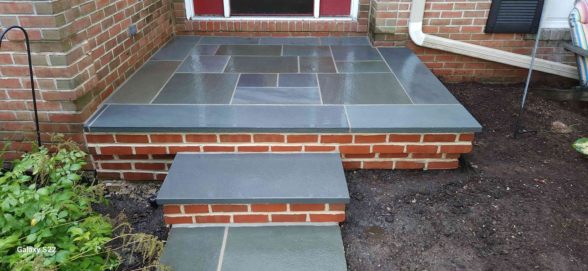 A brick porch with a smooth, dark blue-grey stone top and a single matching step leading up to a house entrance.