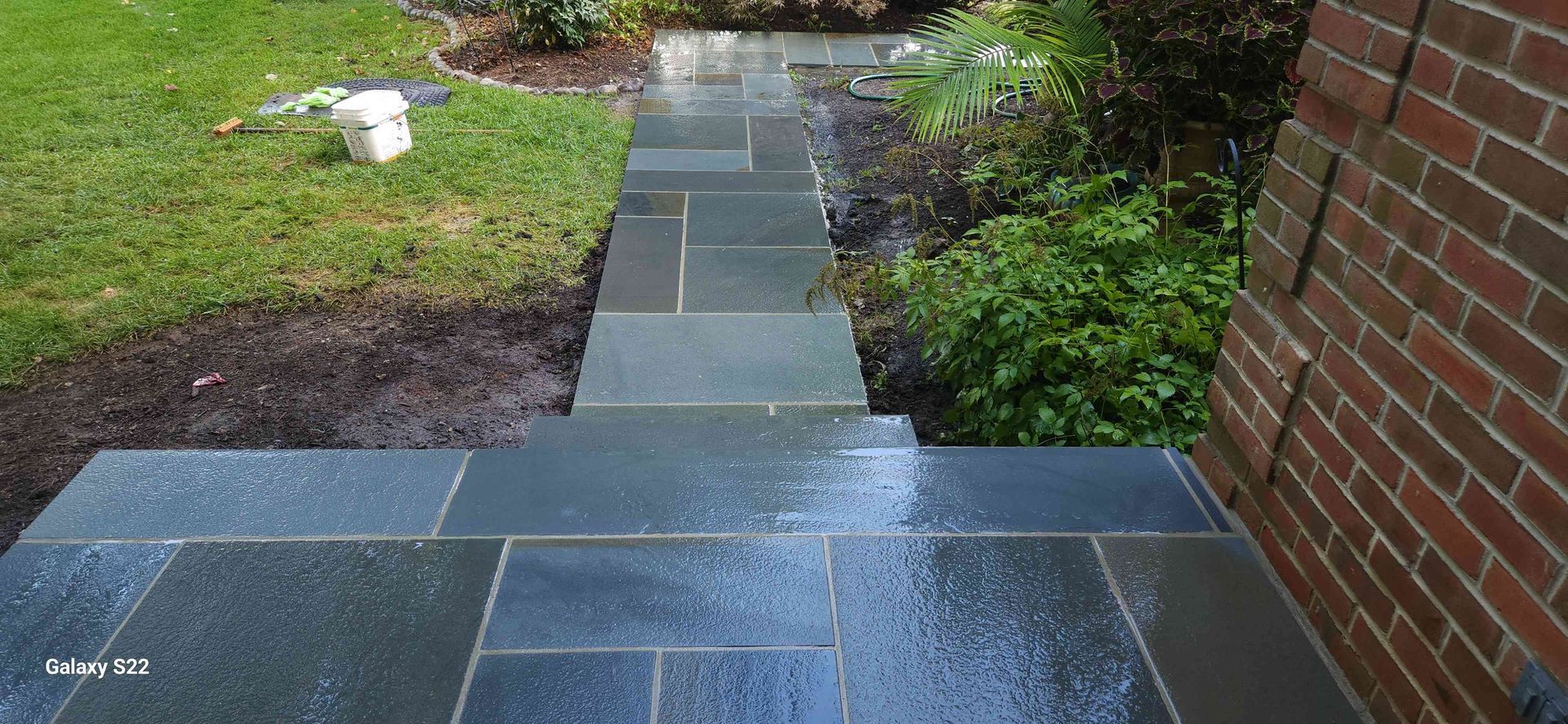 Wet, dark blue stone stairs lead from a brick building wall down to a green lawn.