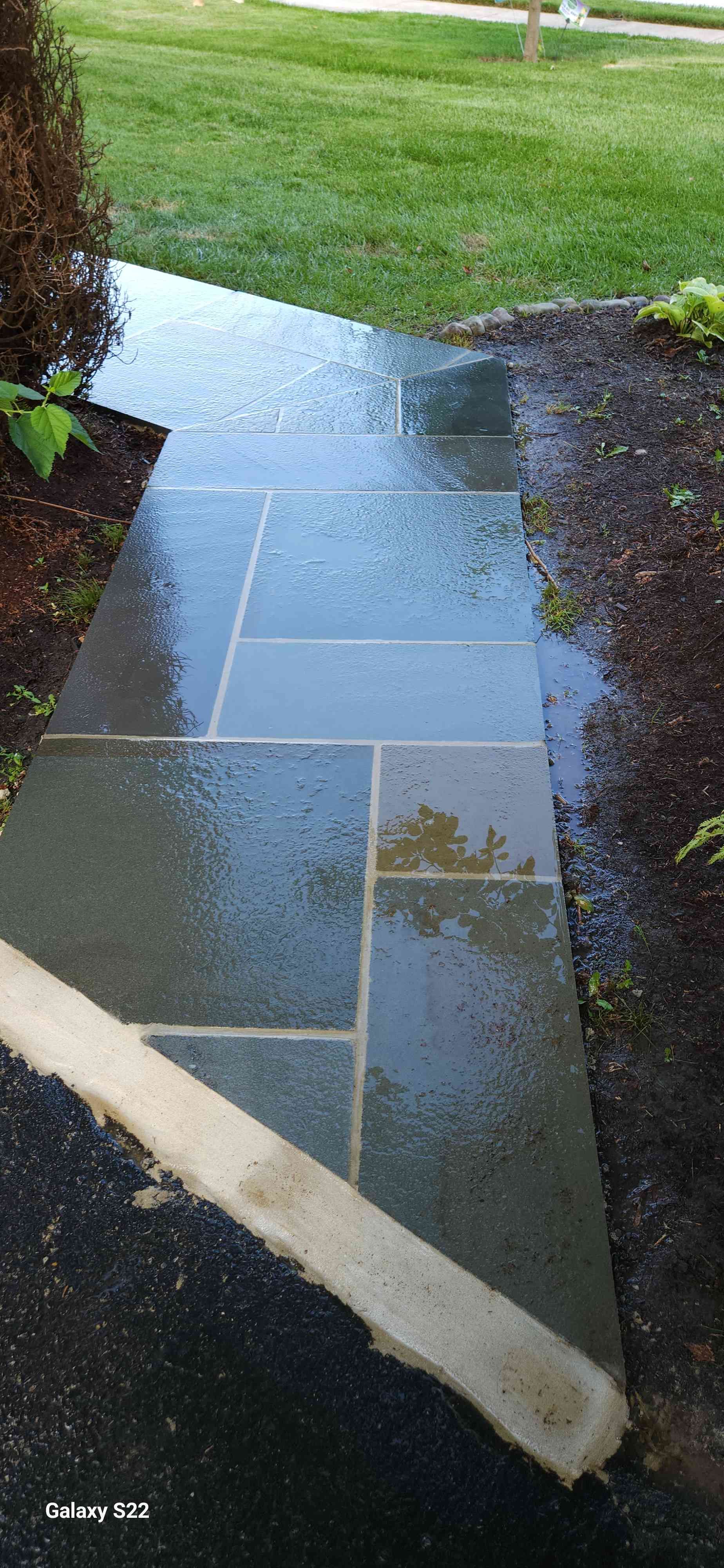 A wet flagstone walkway cuts through a yard with green grass and dark mulch beds.