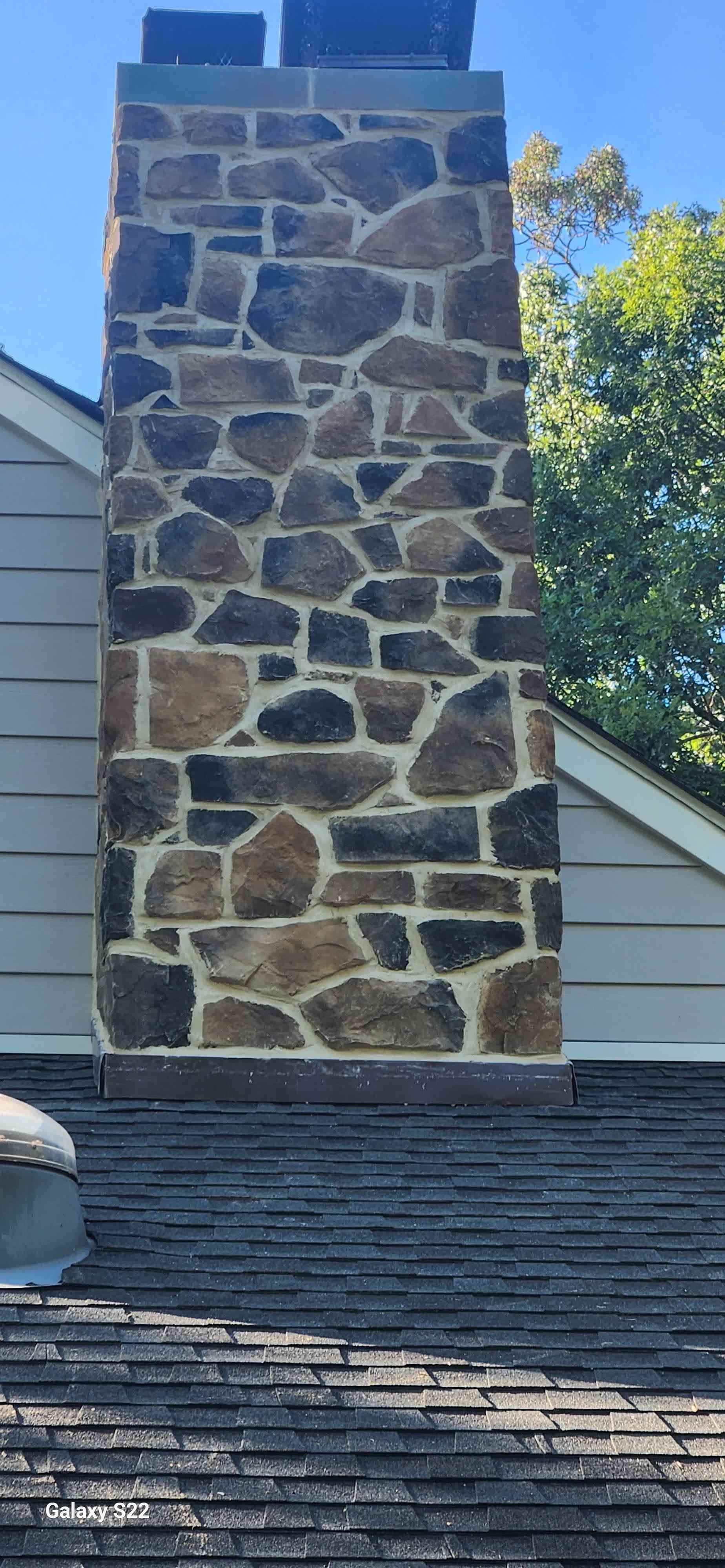 A stone chimney with brown, tan, and grey stones rises from a dark shingled roof against a clear blue sky.