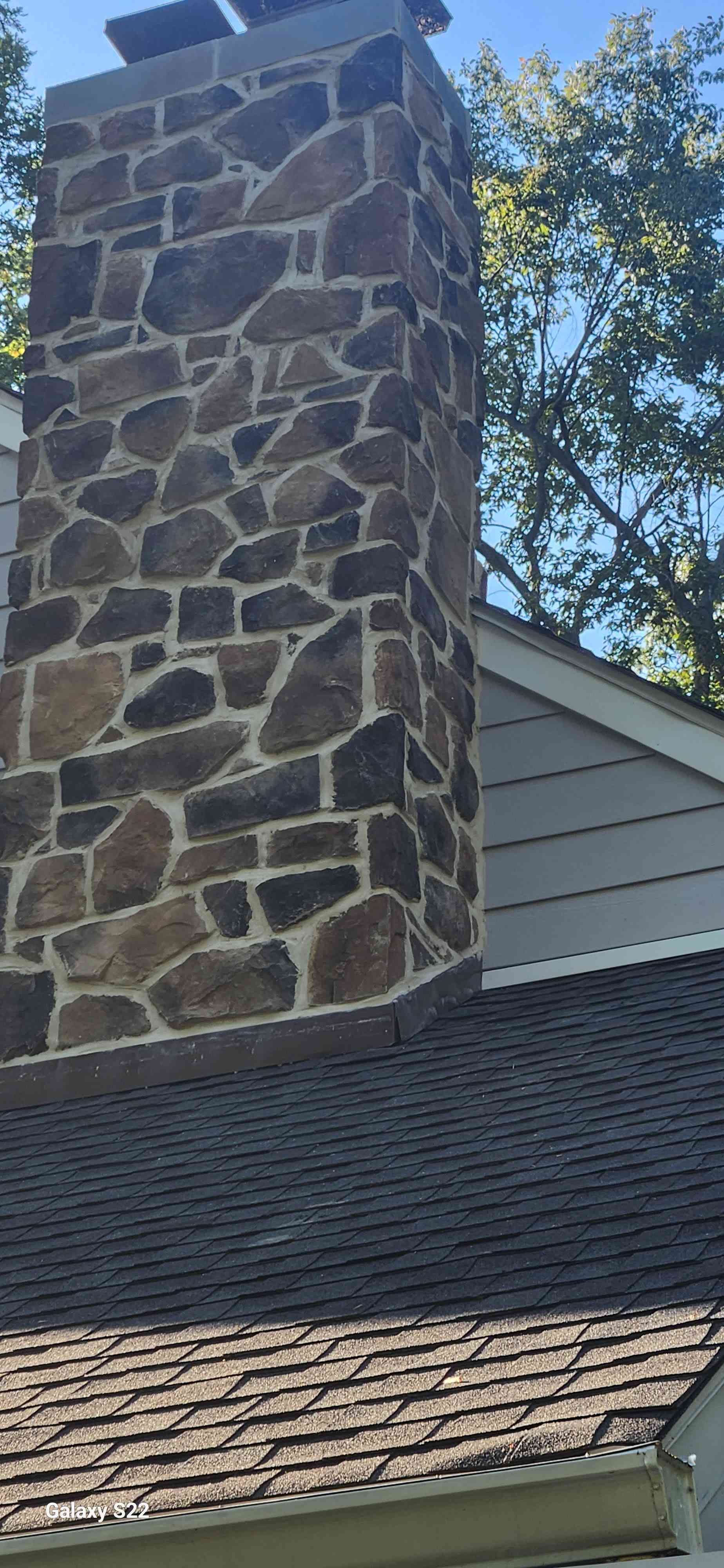 A tall stone chimney with dark, irregular fieldstones rises through the roofline of a house with grey siding.