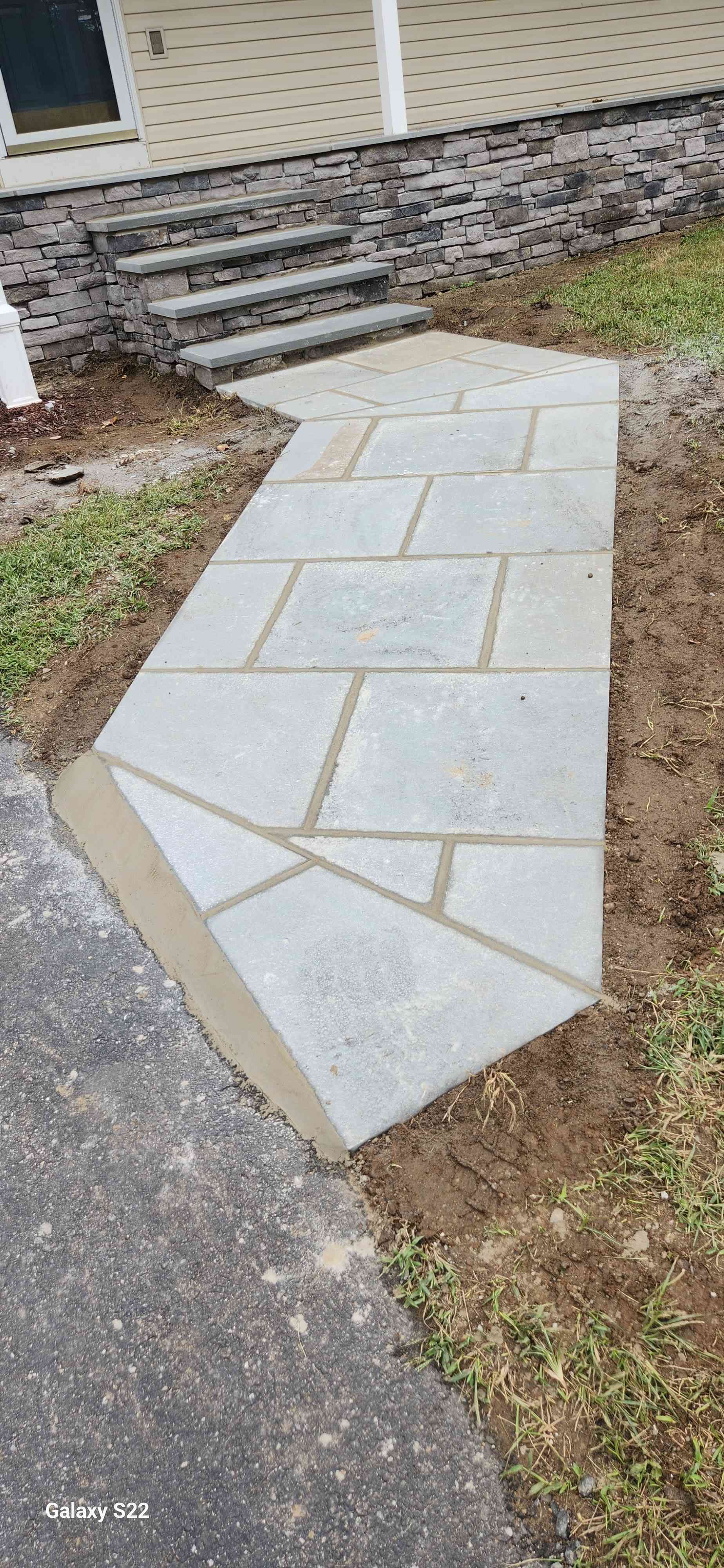 A new light-gray flagstone walkway leading from a paved driveway to a set of stone steps at a house entrance.