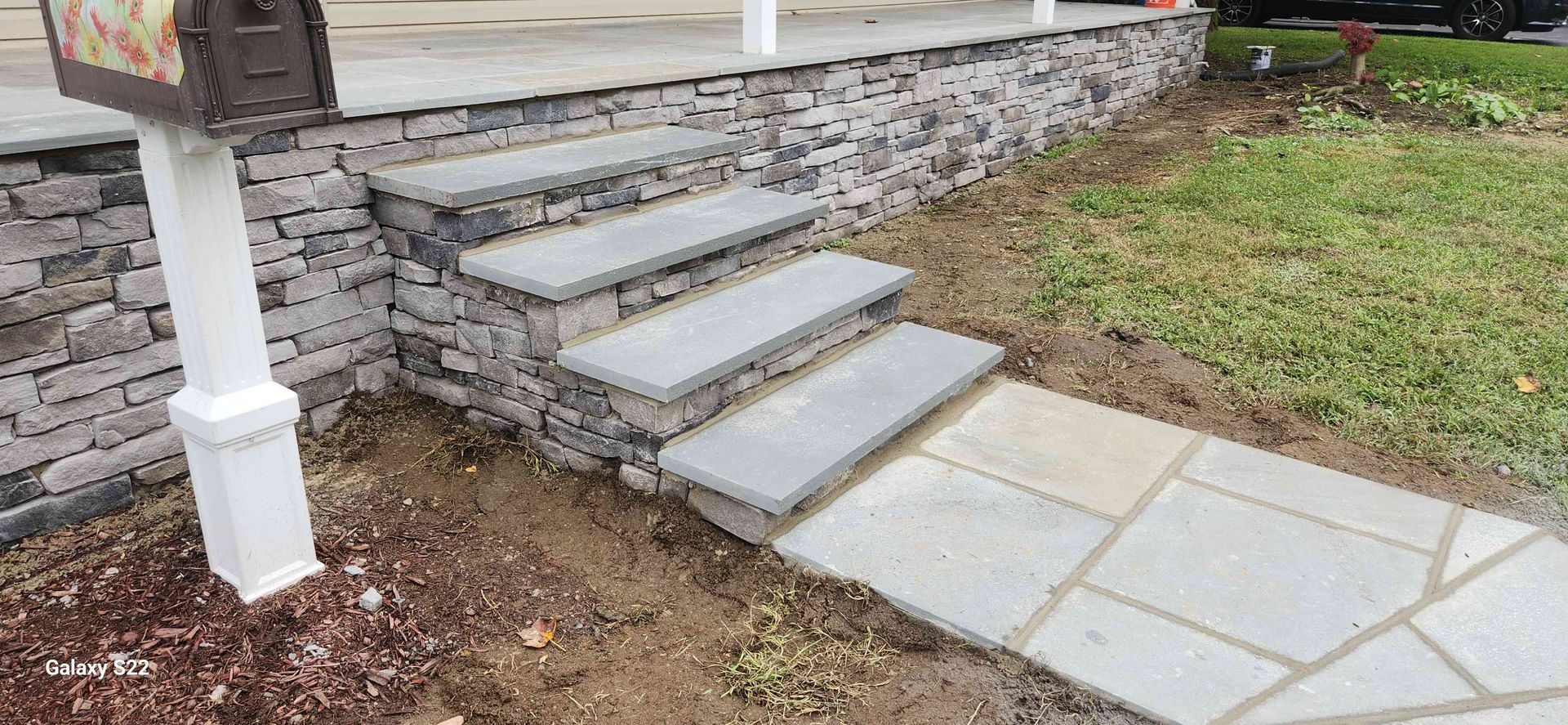 Four stone steps with gray treads leading up to a stone porch, situated next to a mailbox post and stone walkway.
