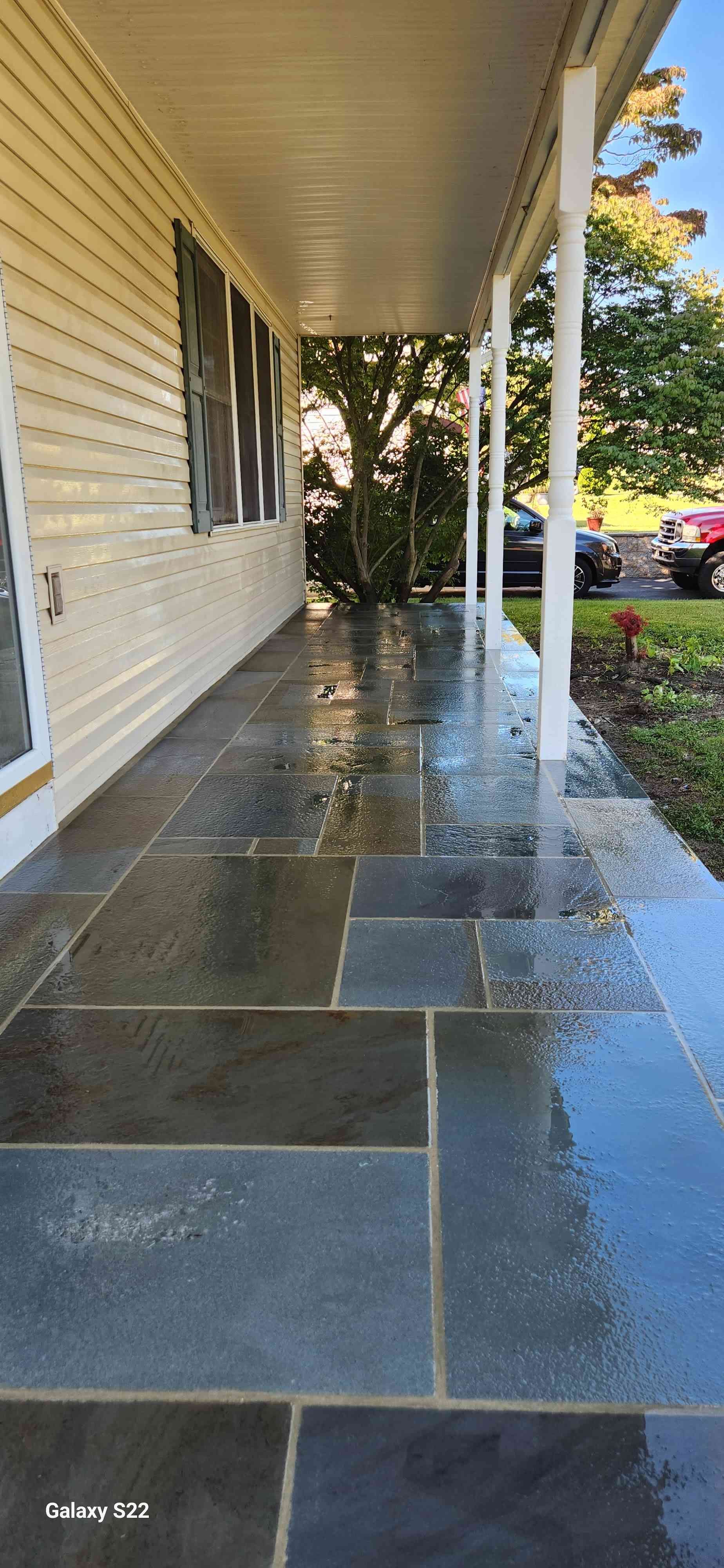 A wet, stone-tiled porch with cream siding, white support columns, and a view of trees and cars in the distance.