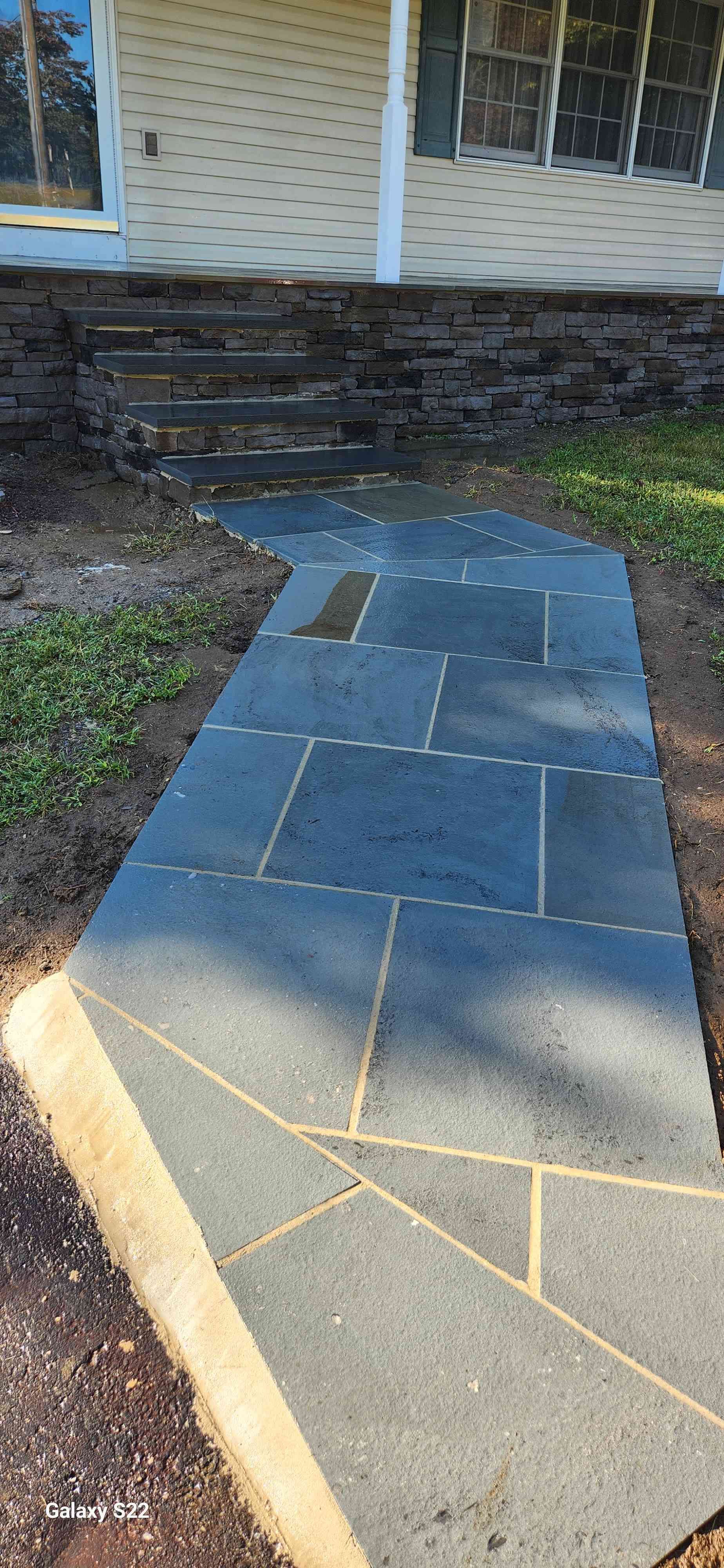 A stone walkway made of large, dark gray, rectangular tiles leads up to the stairs of a house with light-colored siding.