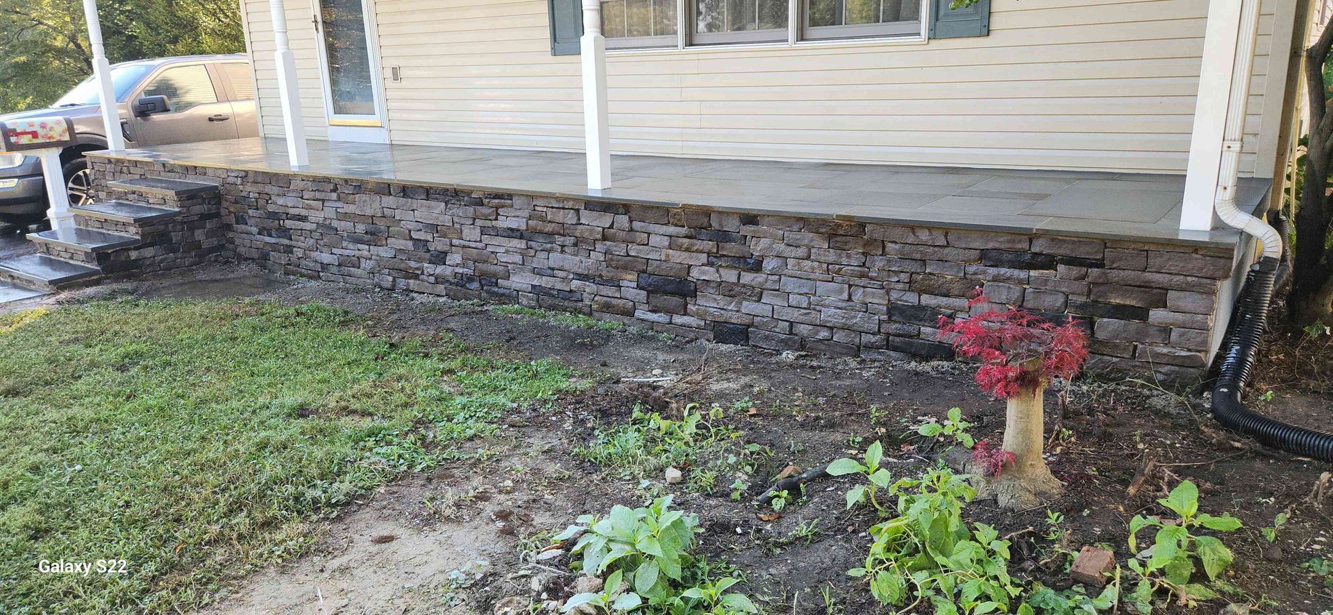 A front porch with a stacked stone foundation, beige siding, and white posts, viewed from a garden bed.