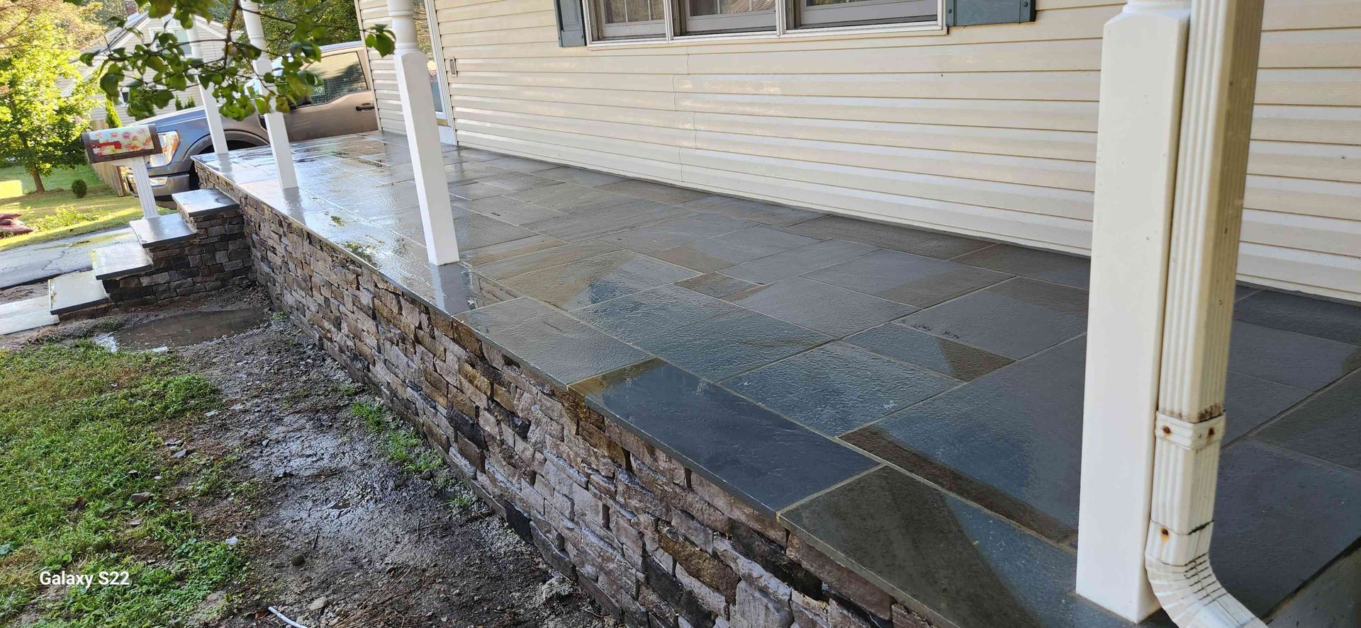 A stone-paved porch with a low brick retaining wall and white vertical posts, seen from an outdoor angle.