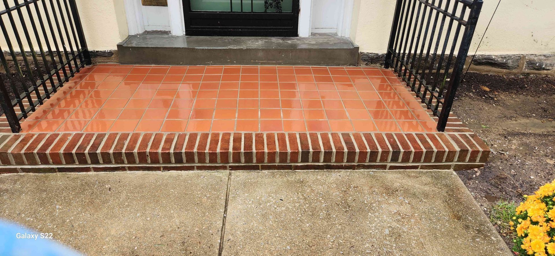 A residential entrance with a rectangular terracotta tile porch bordered by a dark brick edge and black metal railings.