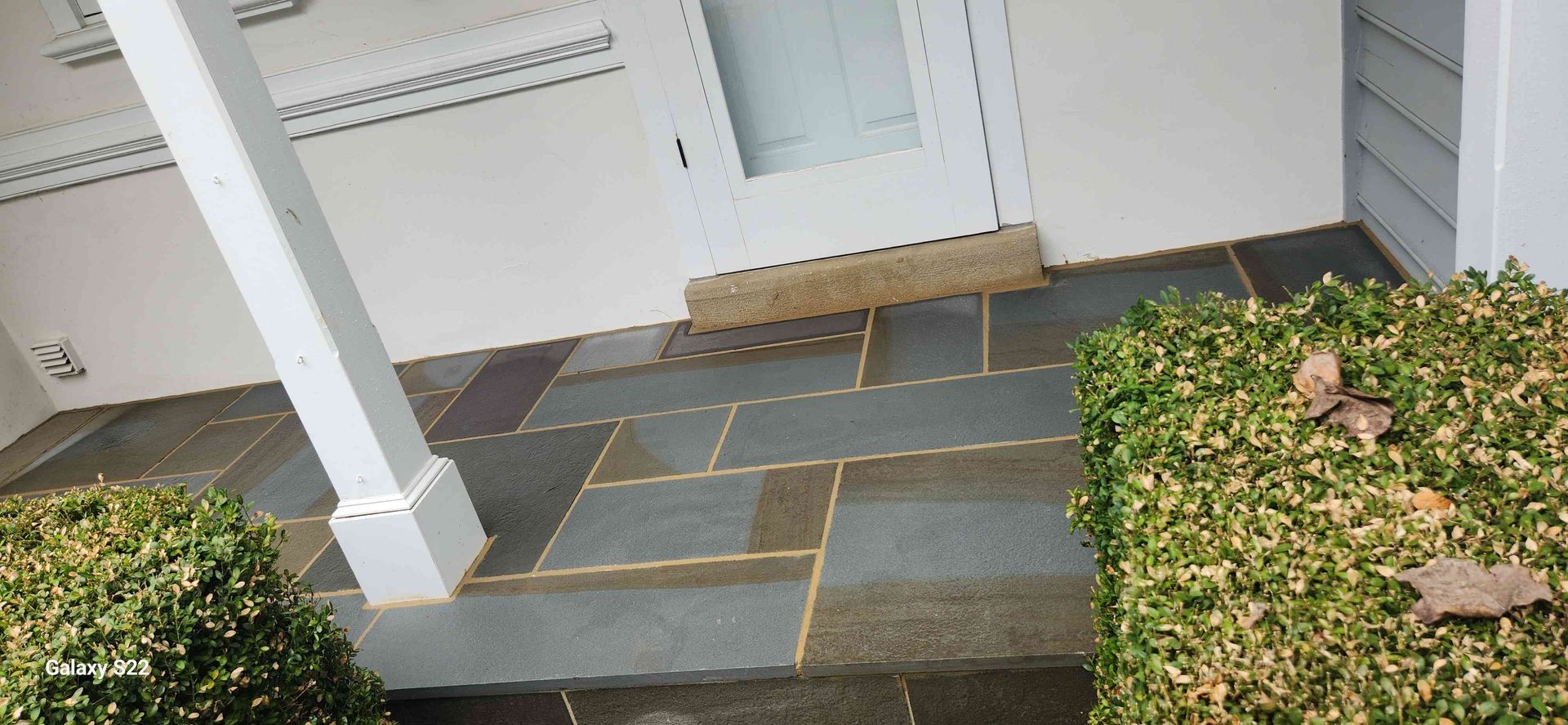 A close-up view of a patio paved with large, rectangular gray stone tiles, featuring two green bushes and a porch pillar.