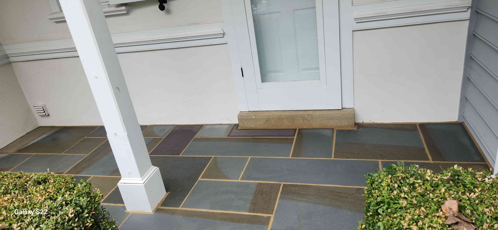A stone patio with a blue-grey, geometric tile pattern leading to a white front door and porch support post.