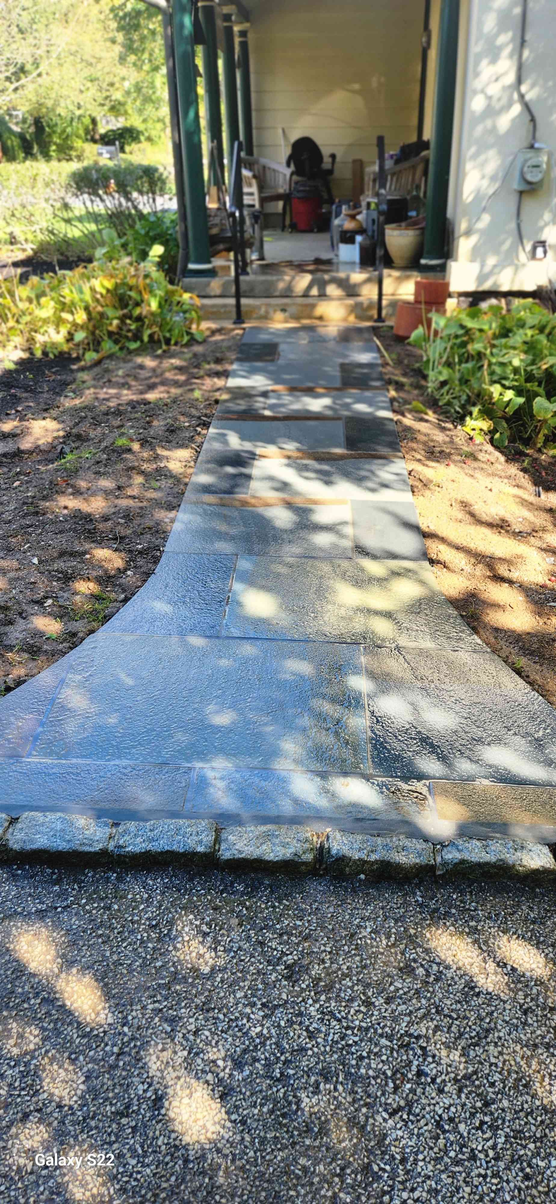 A weathered concrete walkway leads toward a covered porch entrance on a building, surrounded by green landscaping.