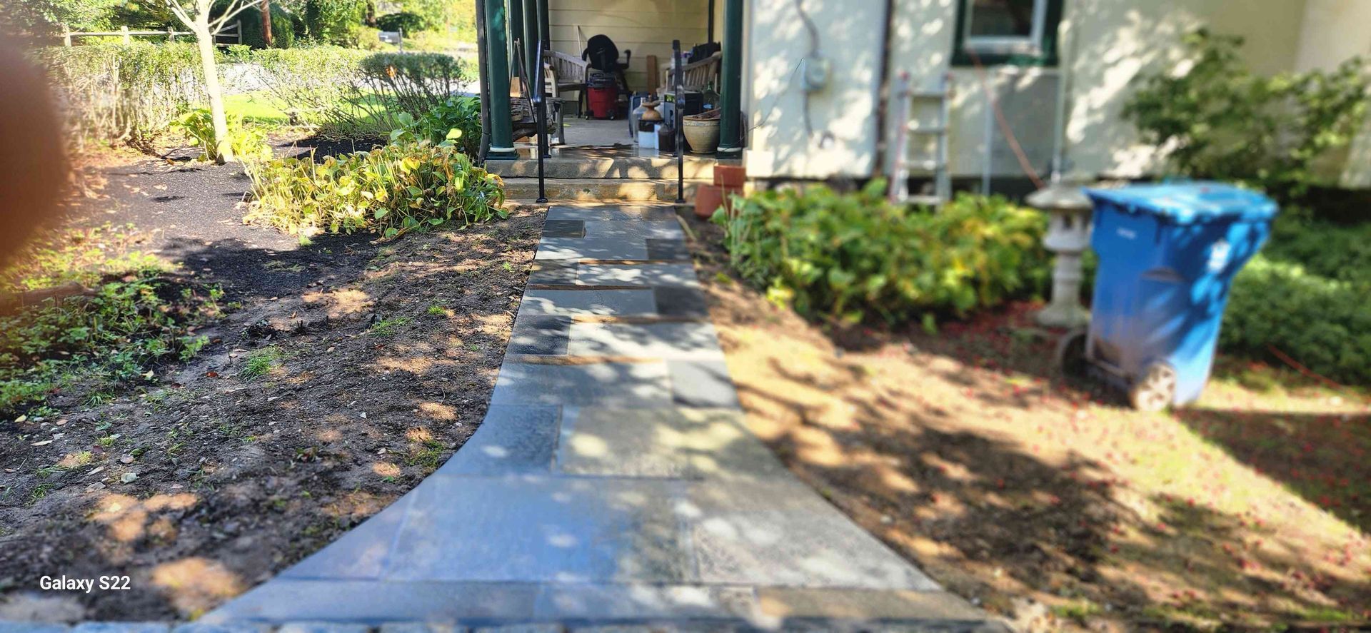 A stone walkway leads to a covered front porch, flanked by mulched garden beds and a blue trash bin on the right.