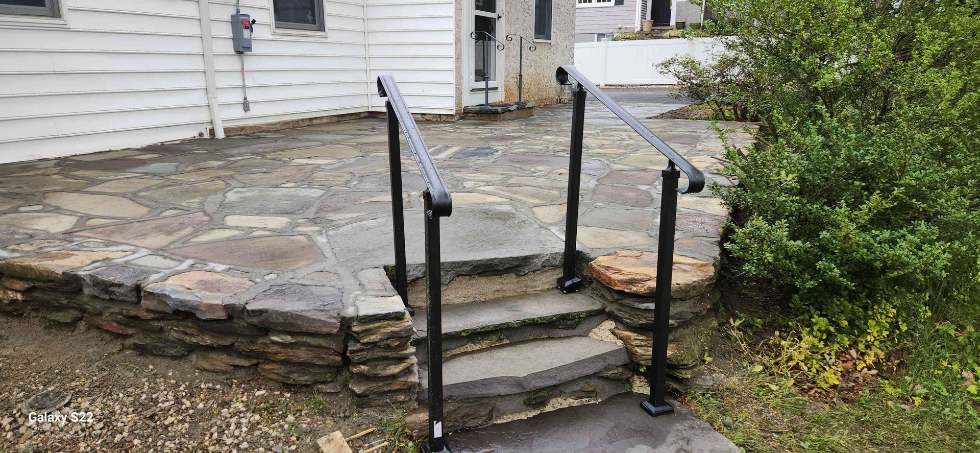 Black metal handrails installed on stone steps leading up to a stone patio next to a white house.