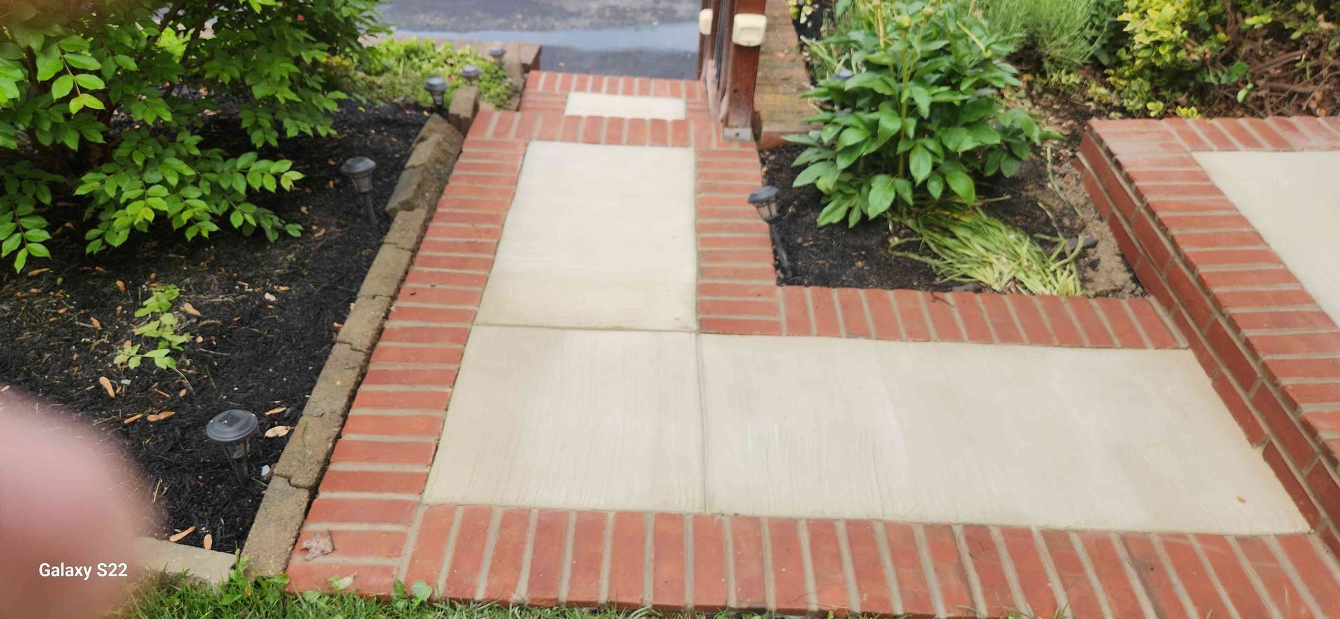 A light-colored stone walkway framed by red bricks, leading toward a house entrance with landscaping on both sides.