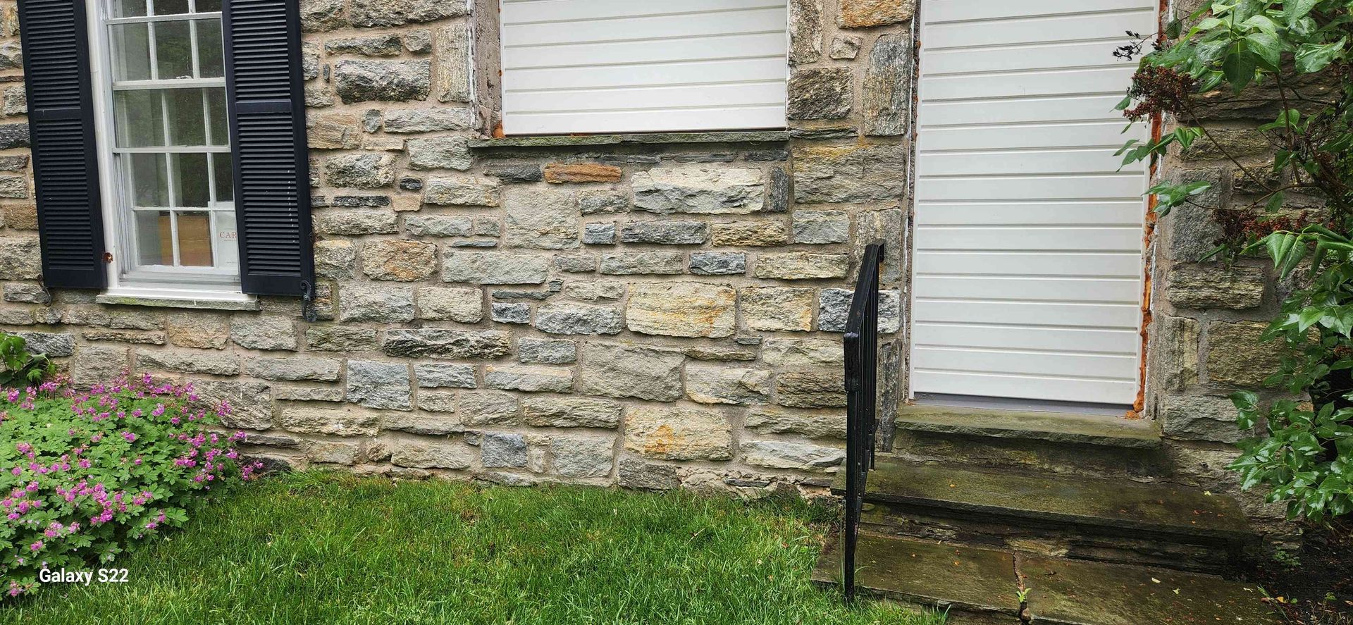 A stone house exterior with a shuttered window, a white door, and steps leading up to an entrance.