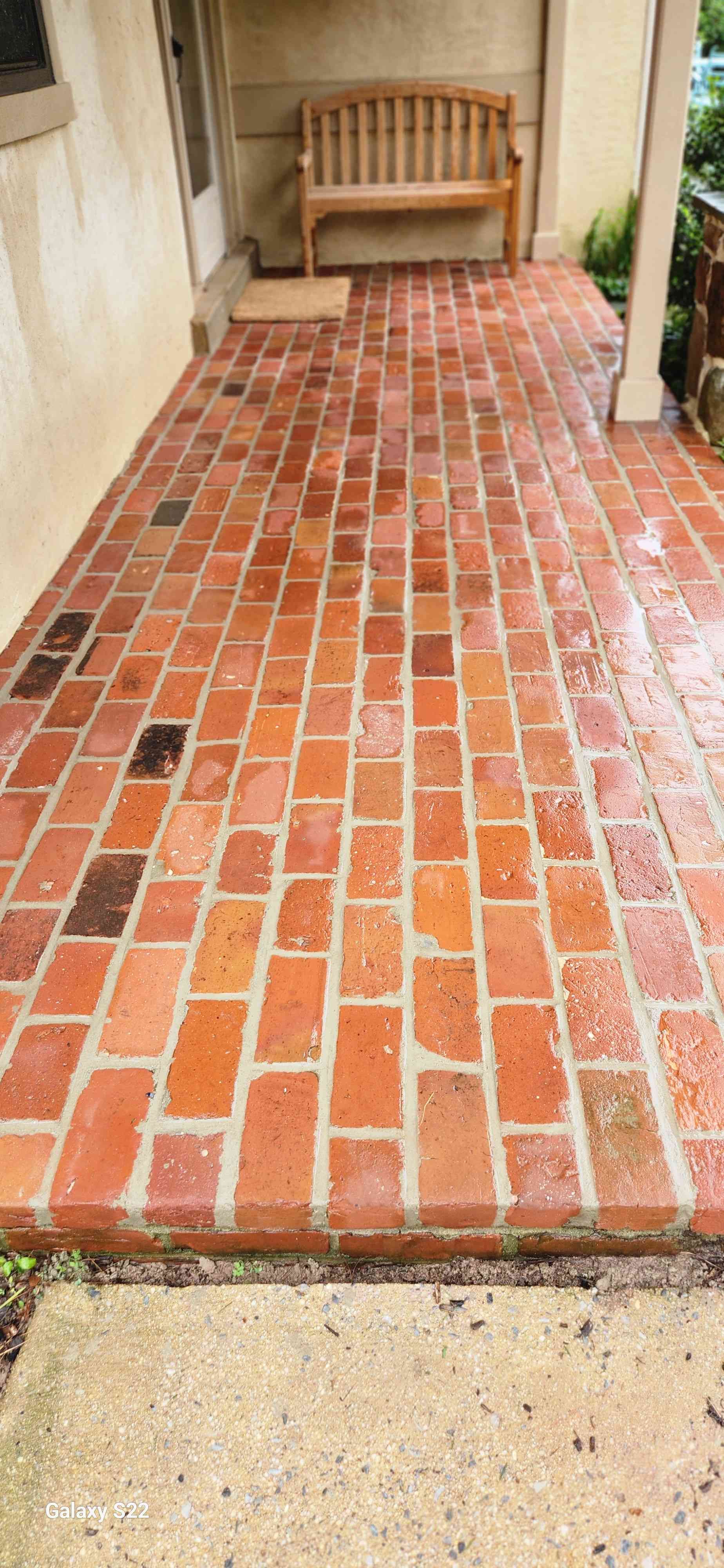 A wooden bench sits on a red brick porch next to a house exterior wall and a support post.