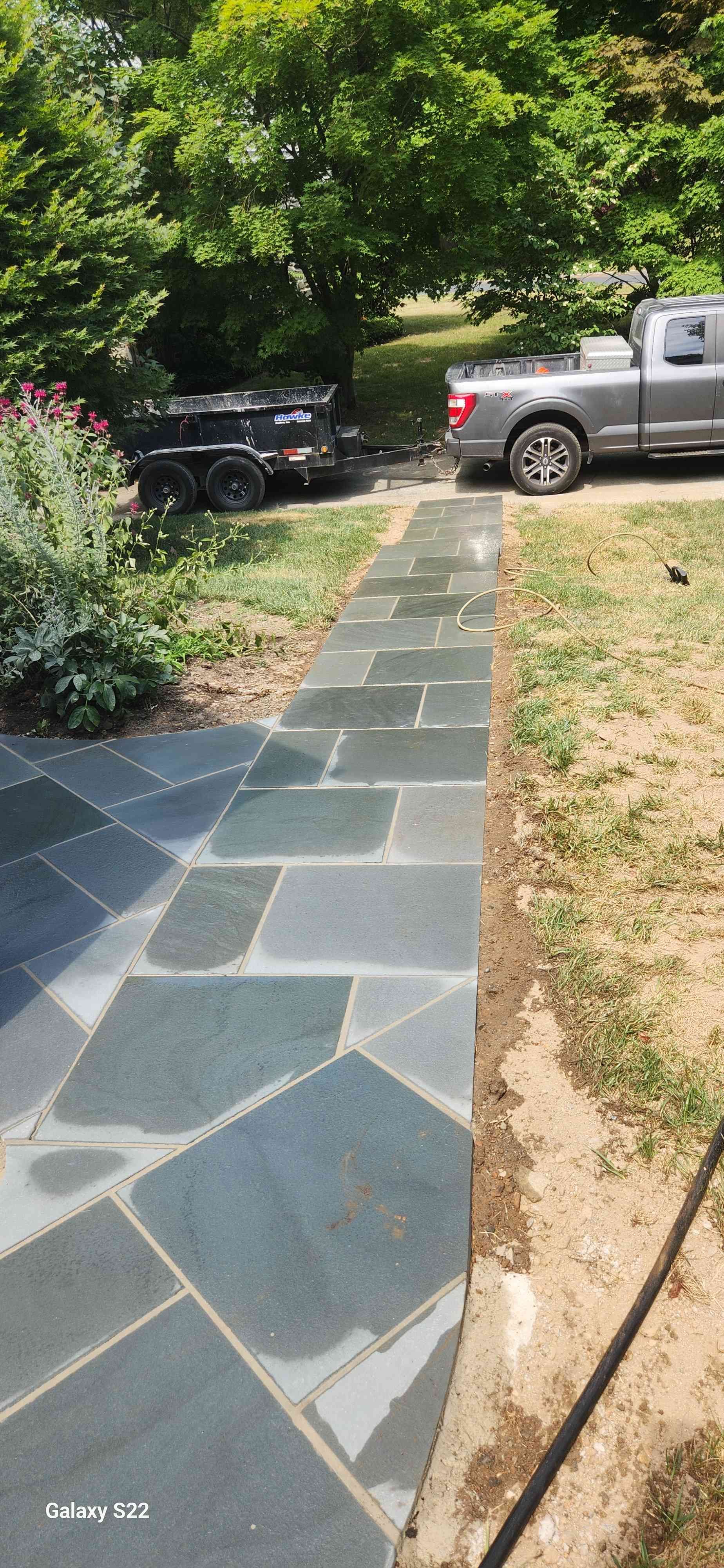 A flagstone walkway made of dark, rectangular pavers stretches through a sunny yard toward a parked truck and trailer.