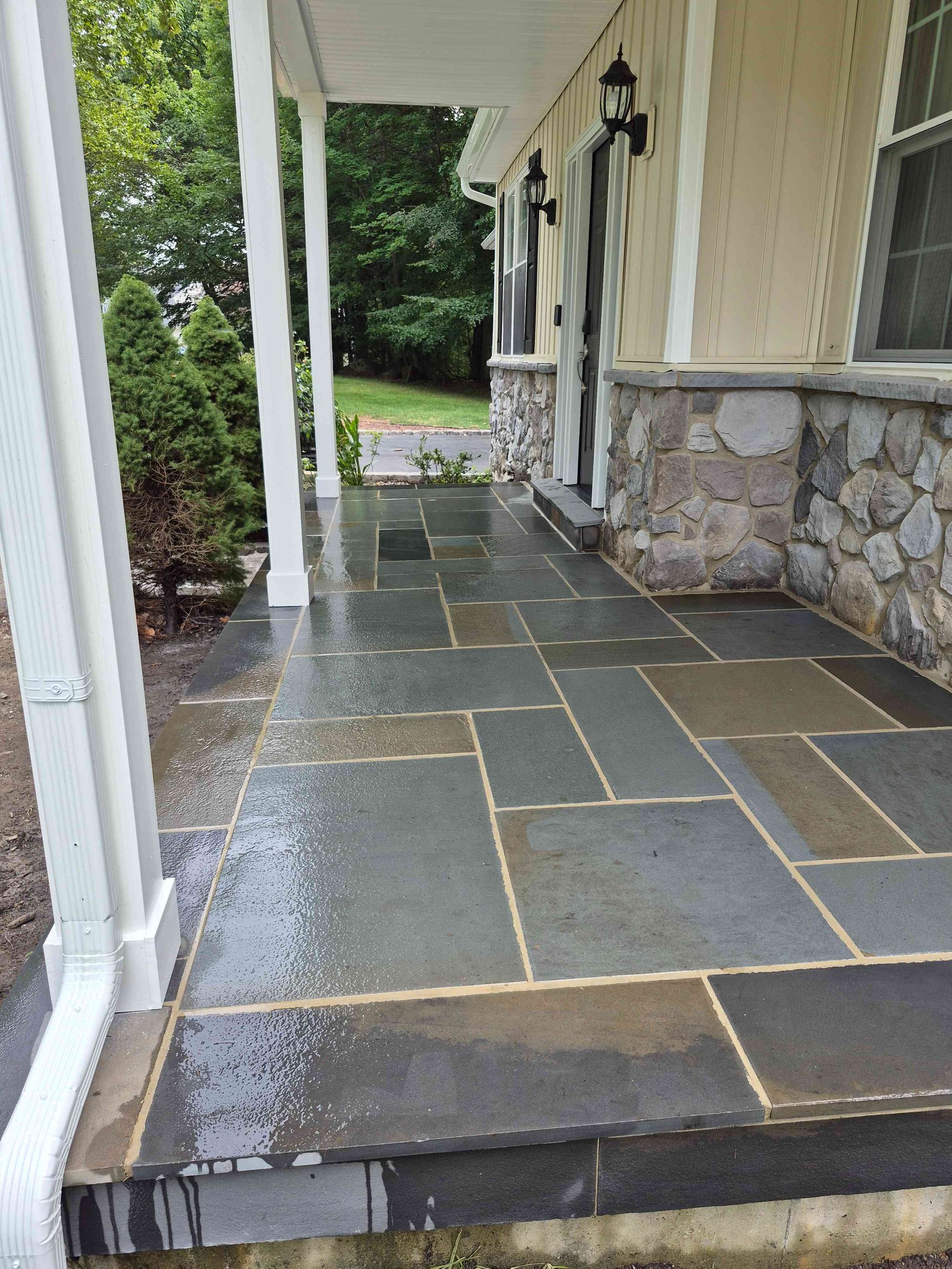 A wet stone porch with rectangular gray and brown tiles, white pillars, and stone siding on a house exterior.