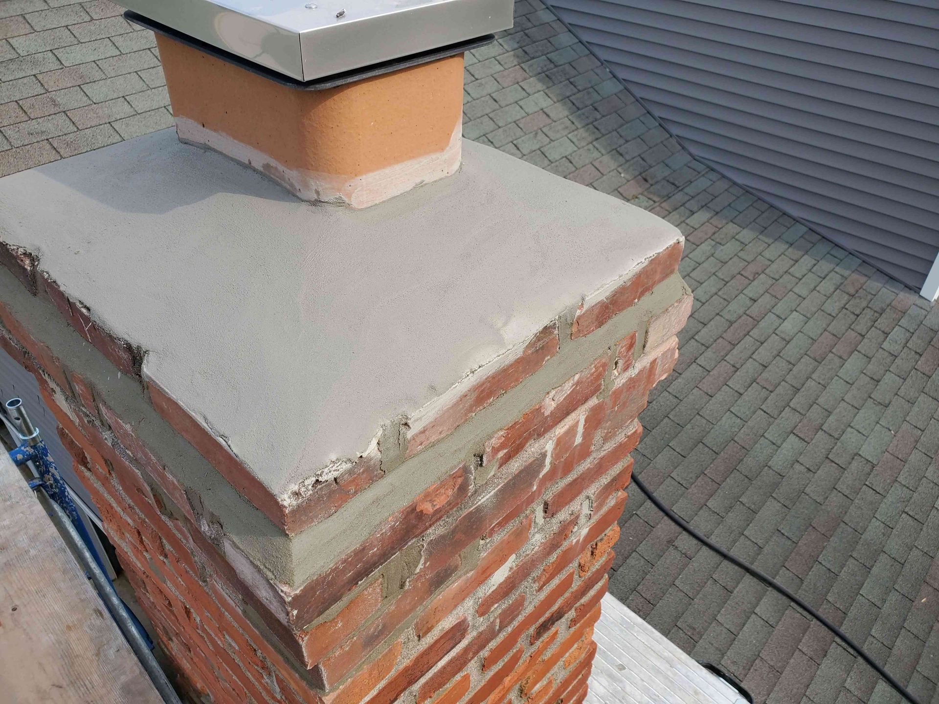 A newly parged concrete chimney crown sits atop a red brick chimney with a metal flue cap, viewed from a roof.