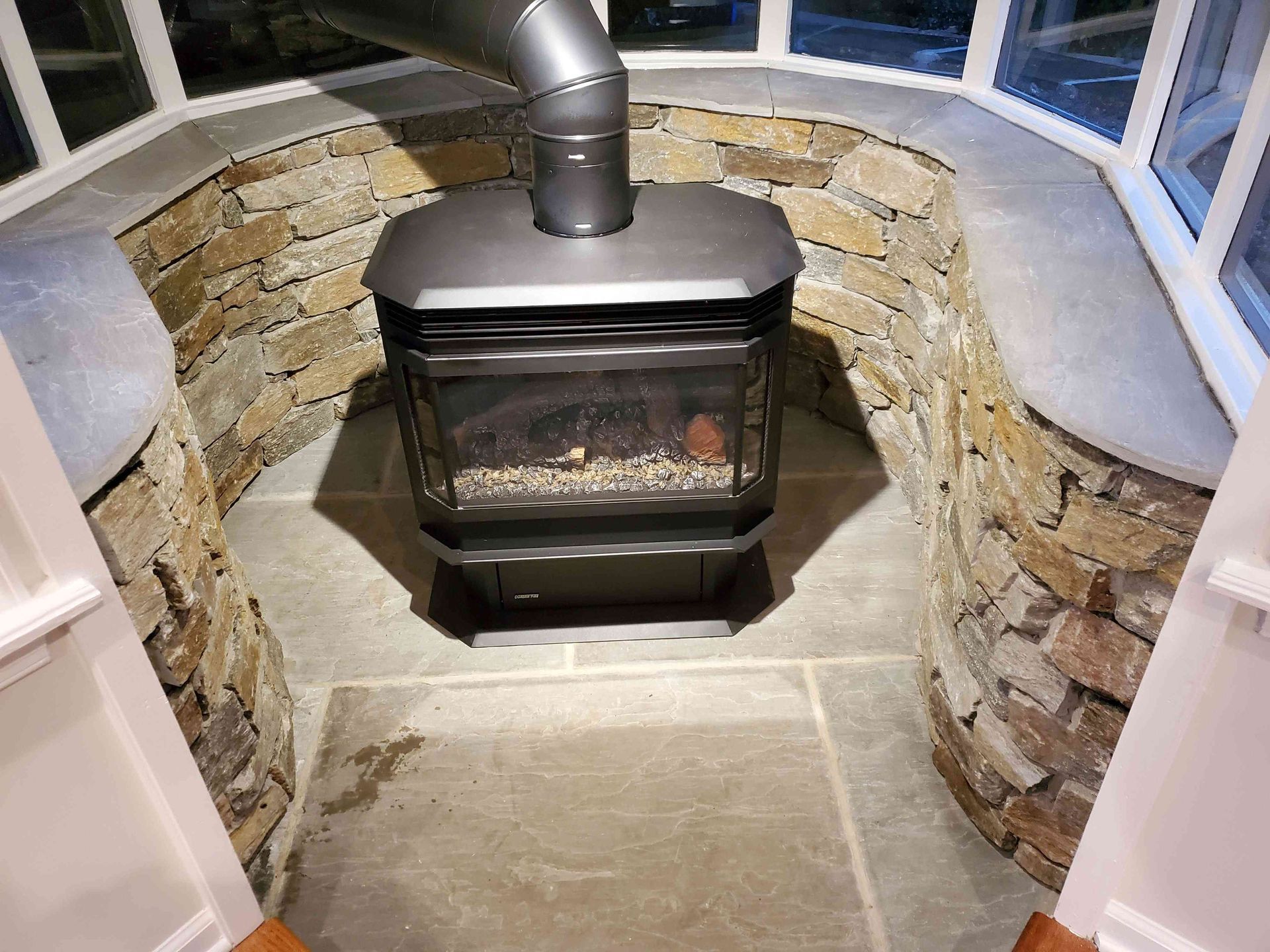 A black wood-burning stove sits on a stone floor in a curved, stone-walled alcove surrounded by large windows.