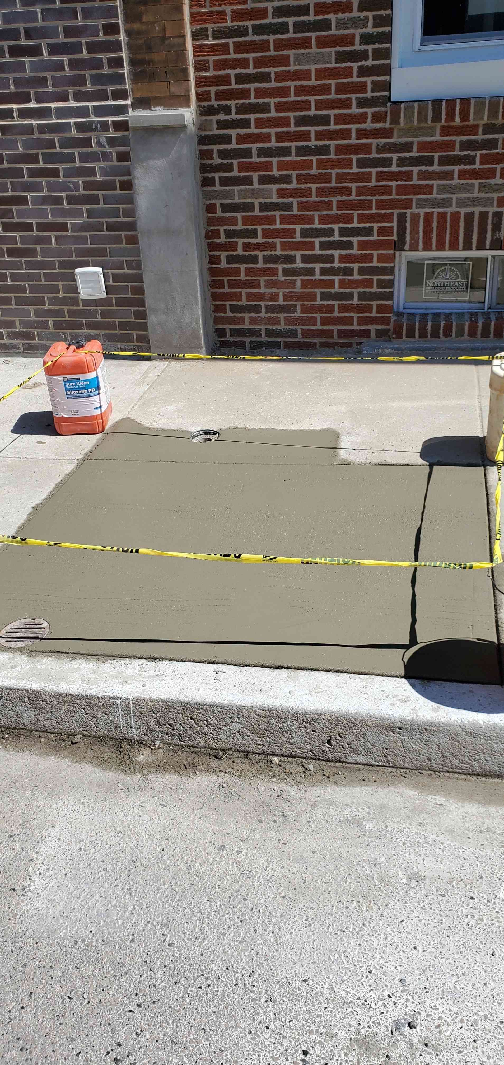 Freshly poured concrete sidewalk section taped off with yellow caution tape in front of a brick building.