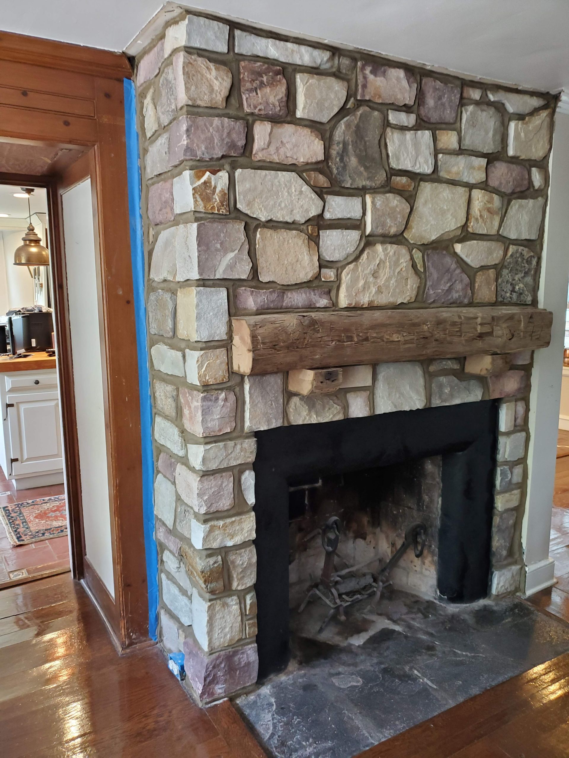 A stone fireplace with a thick wooden mantel stands next to a wooden doorway inside a home.