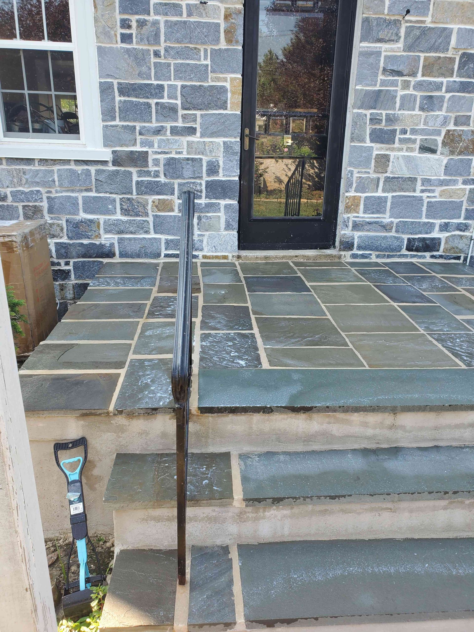 A stone house entryway with blue-gray stone steps and a patio leading to a dark glass door, featuring a black handrail.