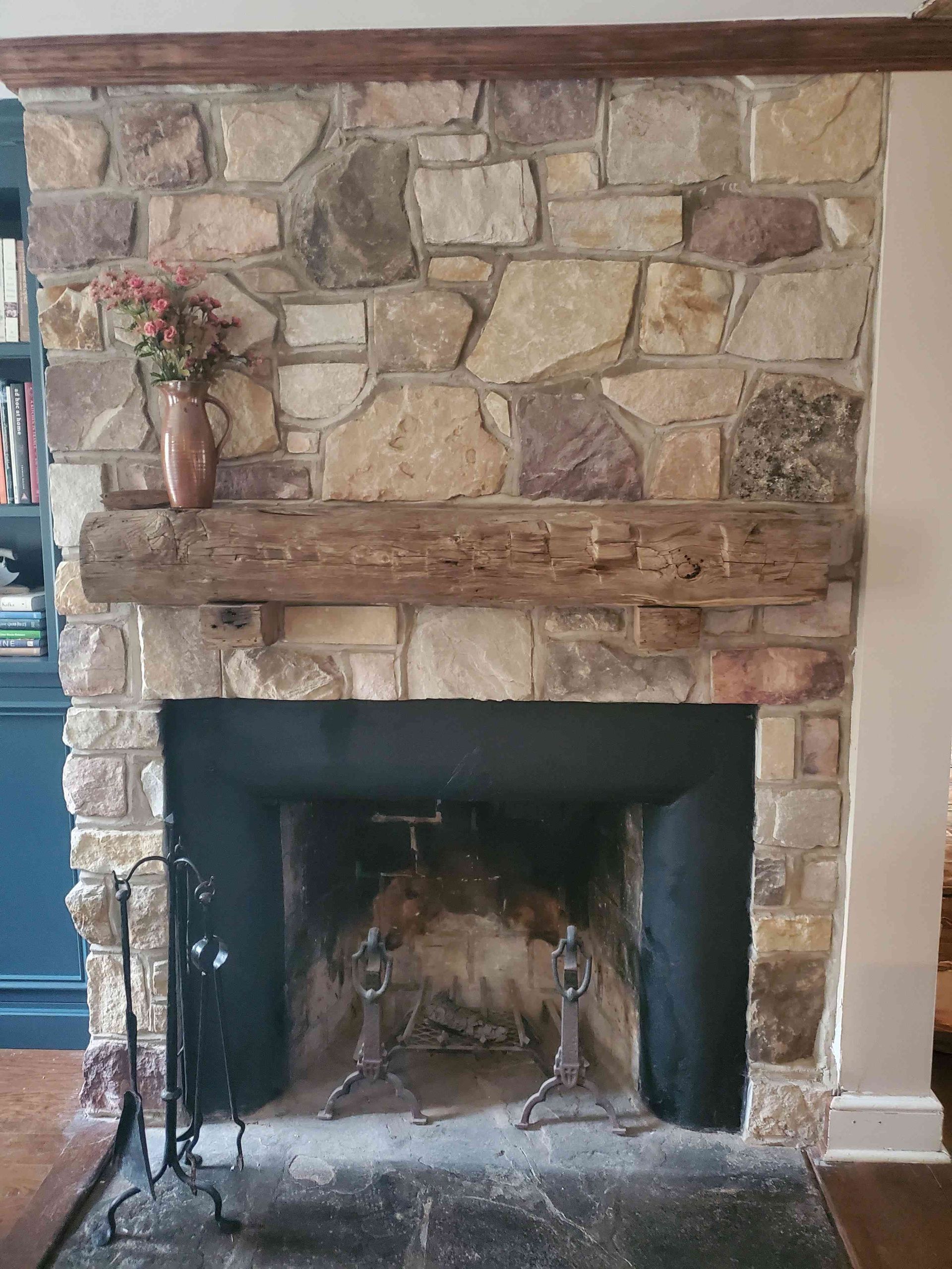 A rustic stone fireplace with a thick, reclaimed wooden mantel and a black metal firebox, accented by a floral vase.