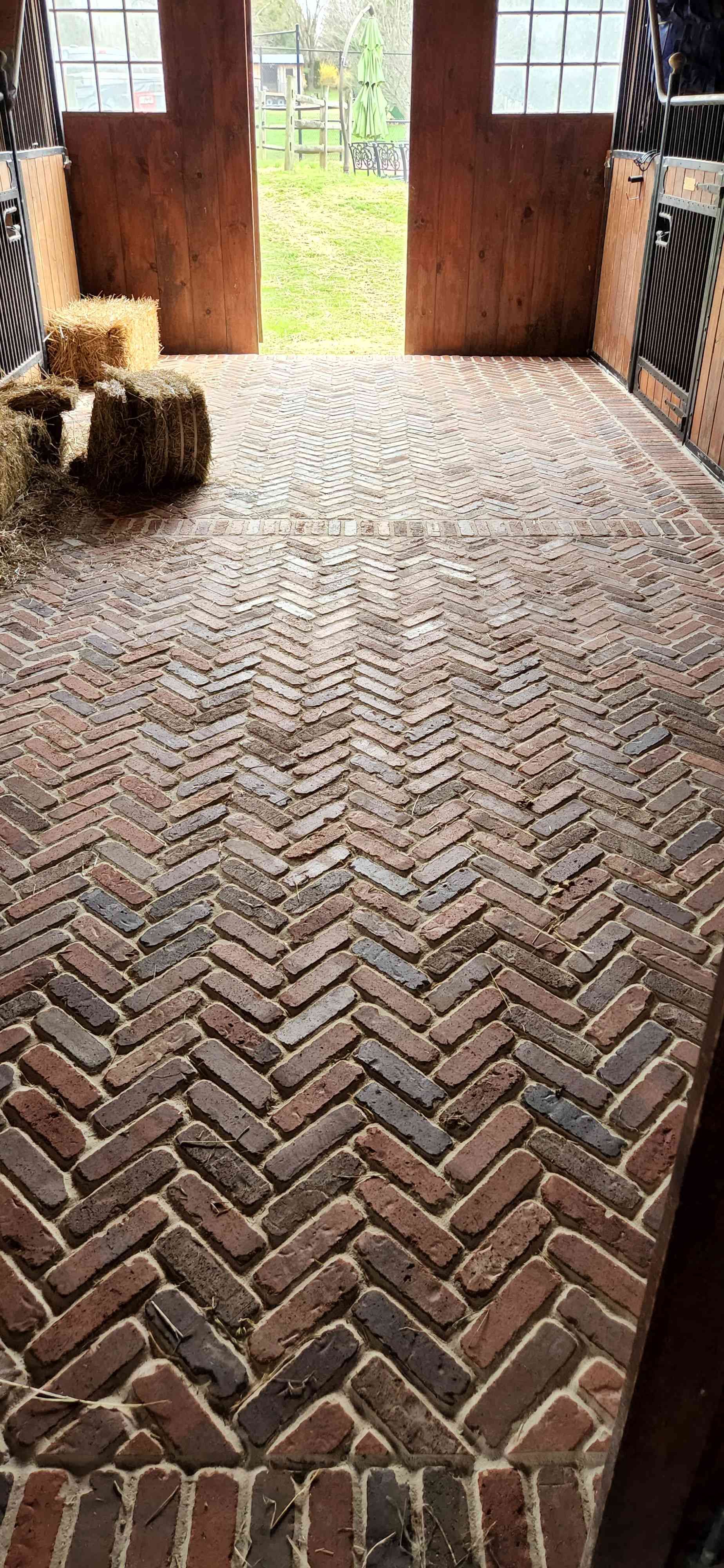A barn floor paved with red and brown bricks in a herringbone pattern, leading to open double doors looking outdoors.
