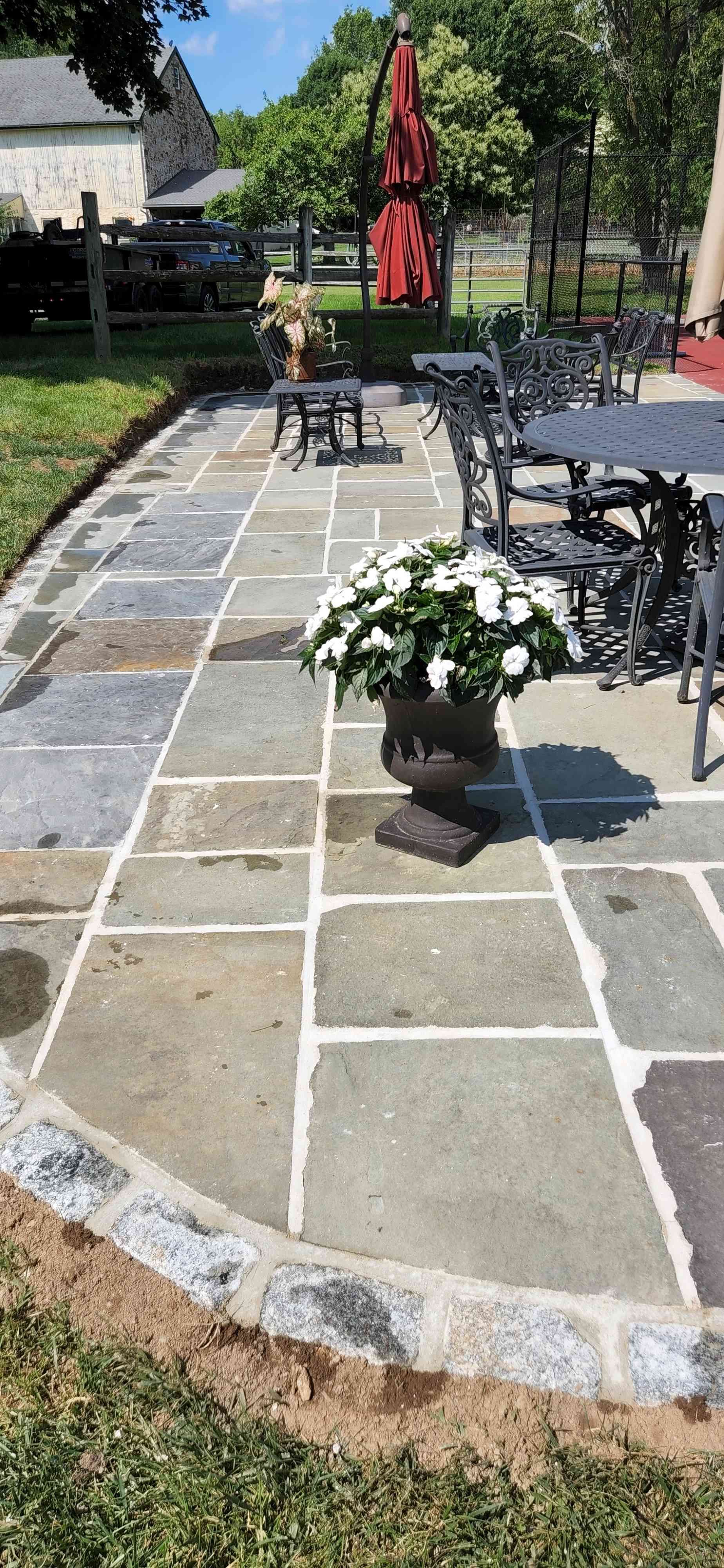A stone patio with a potted white flower arrangement, wrought iron furniture, and a red patio umbrella in a garden.