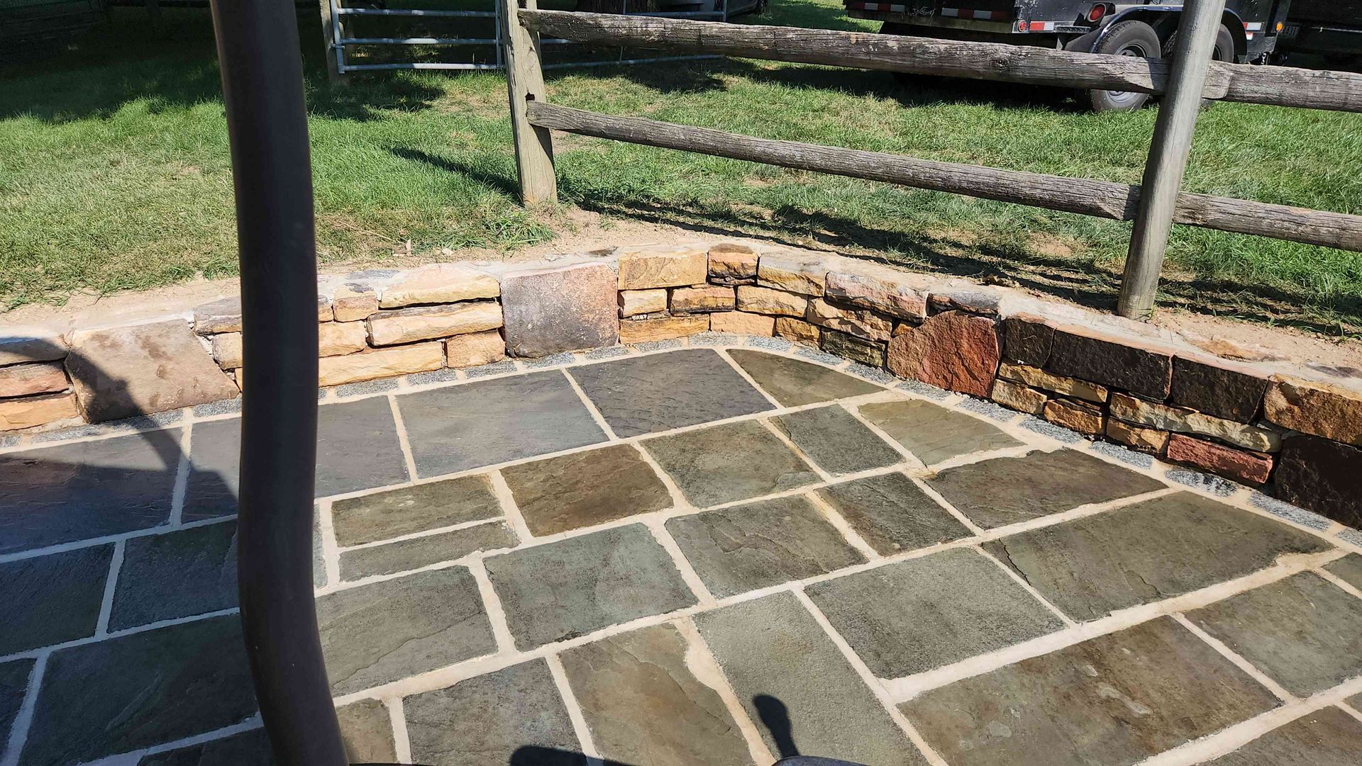 A stone patio with a low, decorative brick wall border in a grassy yard, partially shaded by a pole.