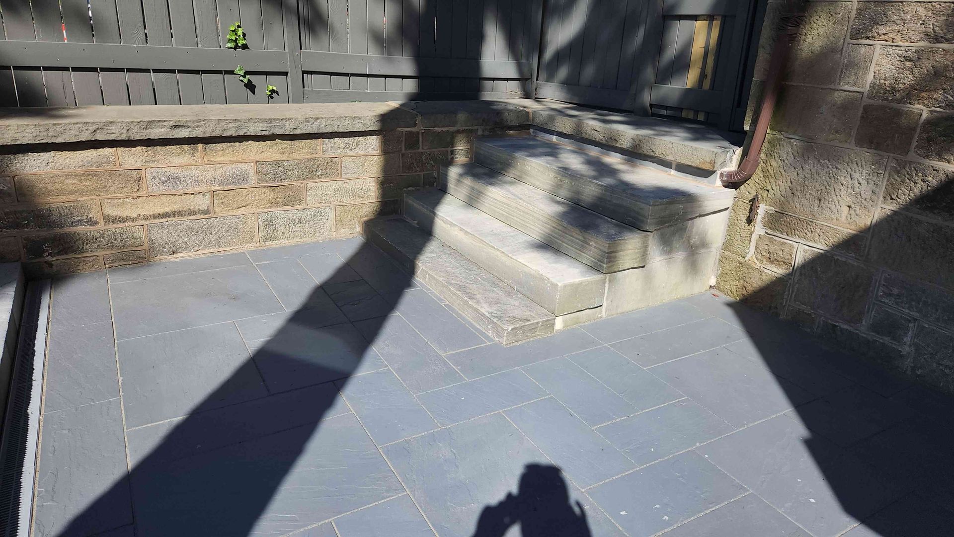 A stone patio with steps leading to a slightly elevated area, bordered by a low stone wall and a grey wooden fence.