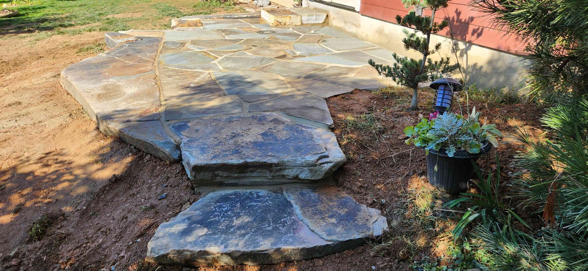 A stone patio and walkway steps set in a yard next to a red building with mulch and plants nearby.