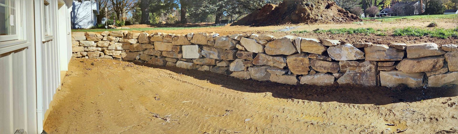A low stone retaining wall curves through a dirt yard next to the side of a white building on a sunny day.