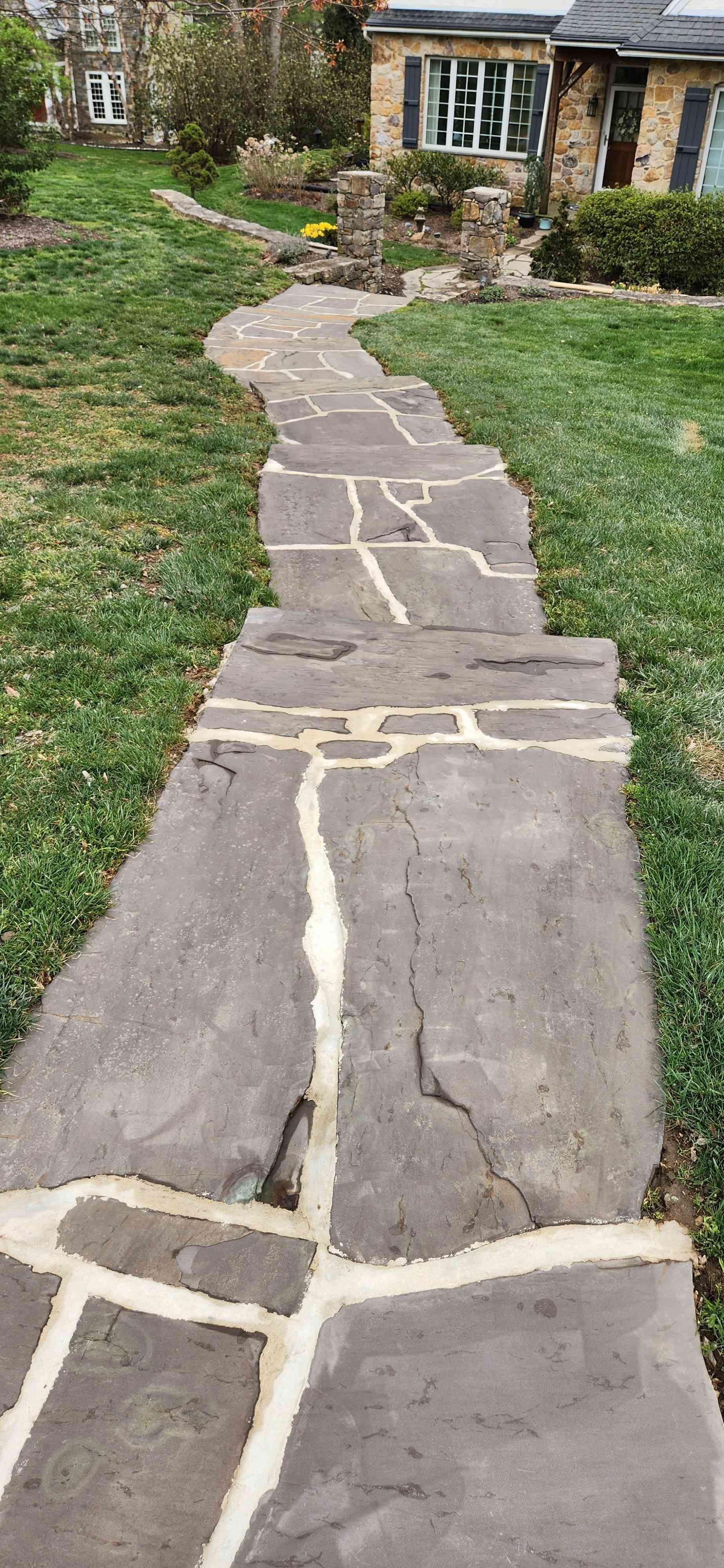 A gray stone walkway with thick, light-colored grout paths leads through a green lawn to a stone house.