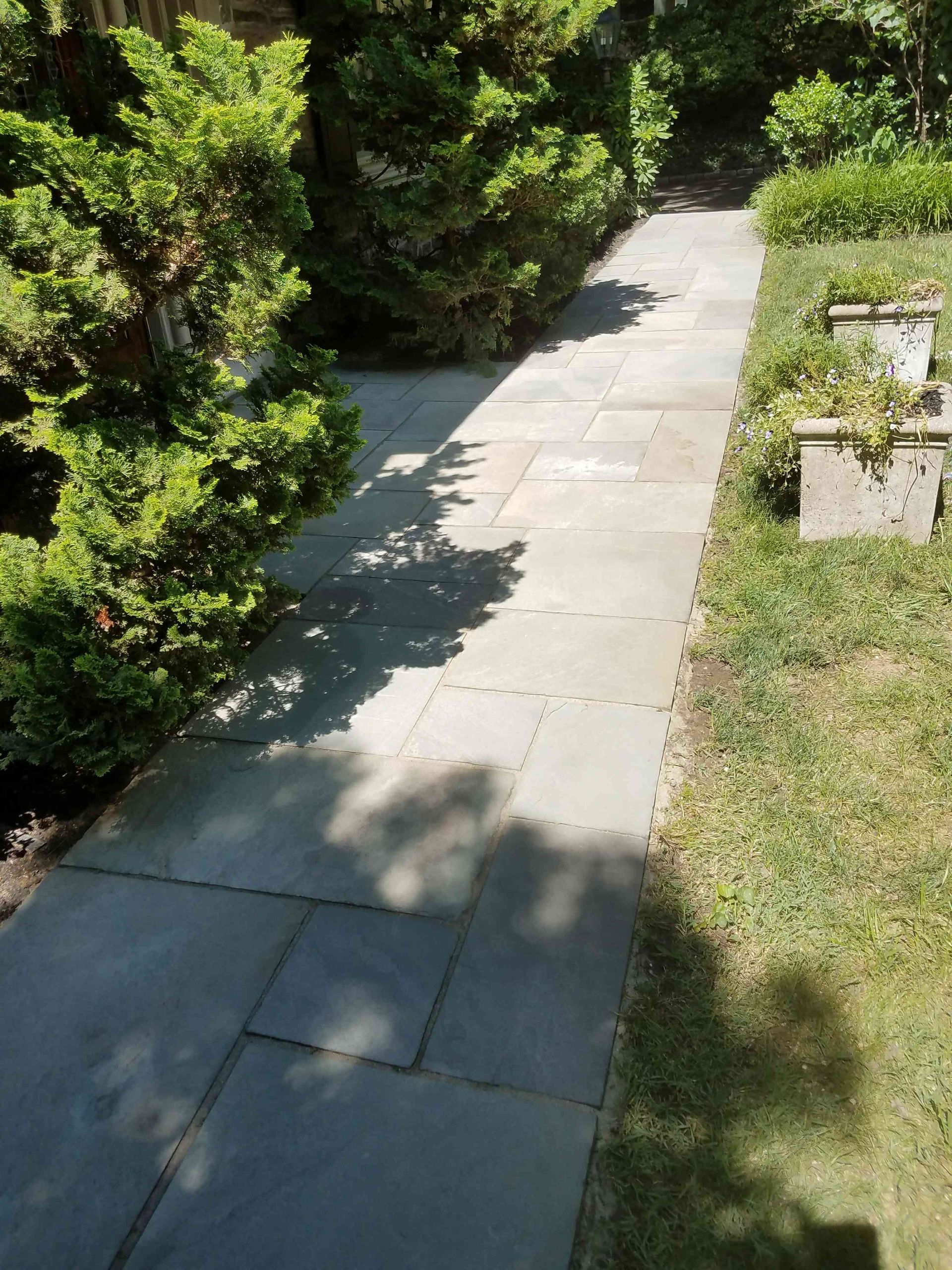 A stone pathway stretches through a garden, bordered by green shrubs on the left and a grass lawn with stone planters.