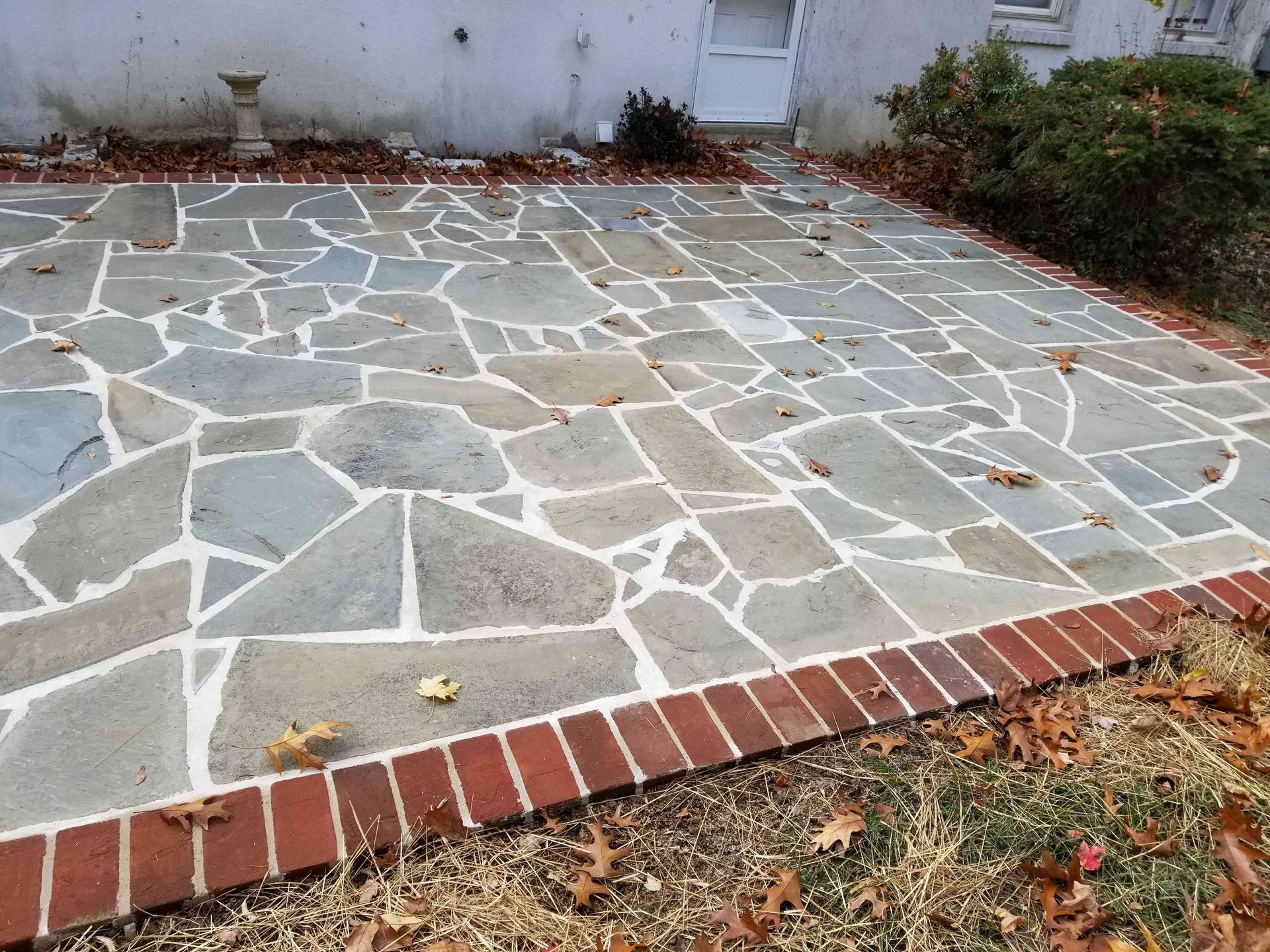 Flagstone patio with a red brick border, set against a house exterior with scattered autumn leaves.