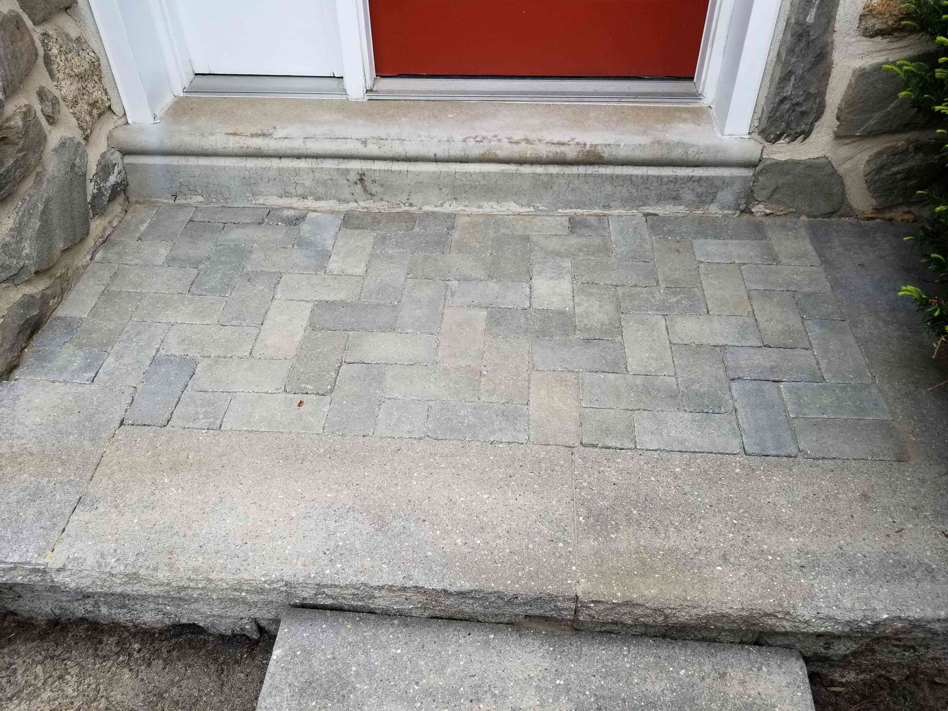 A stone porch with a herringbone paver inlay sits before a red door and white frame, flanked by stone walls.