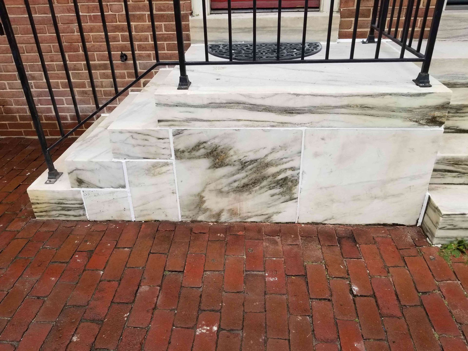 A white marble porch with a black metal railing above a red brick walkway.