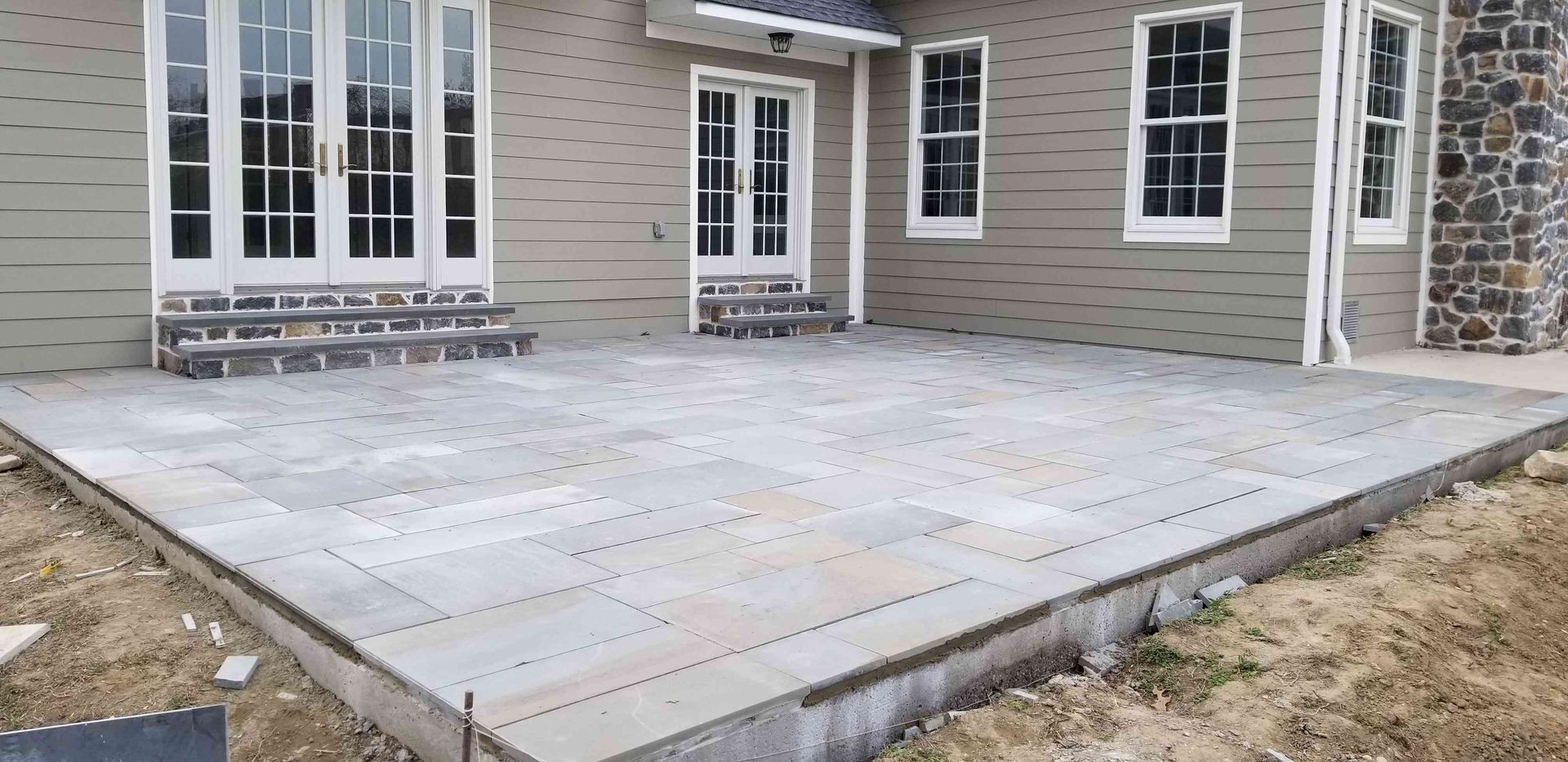 A light-colored stone patio installed outside the back of a house with two sets of French doors and siding.