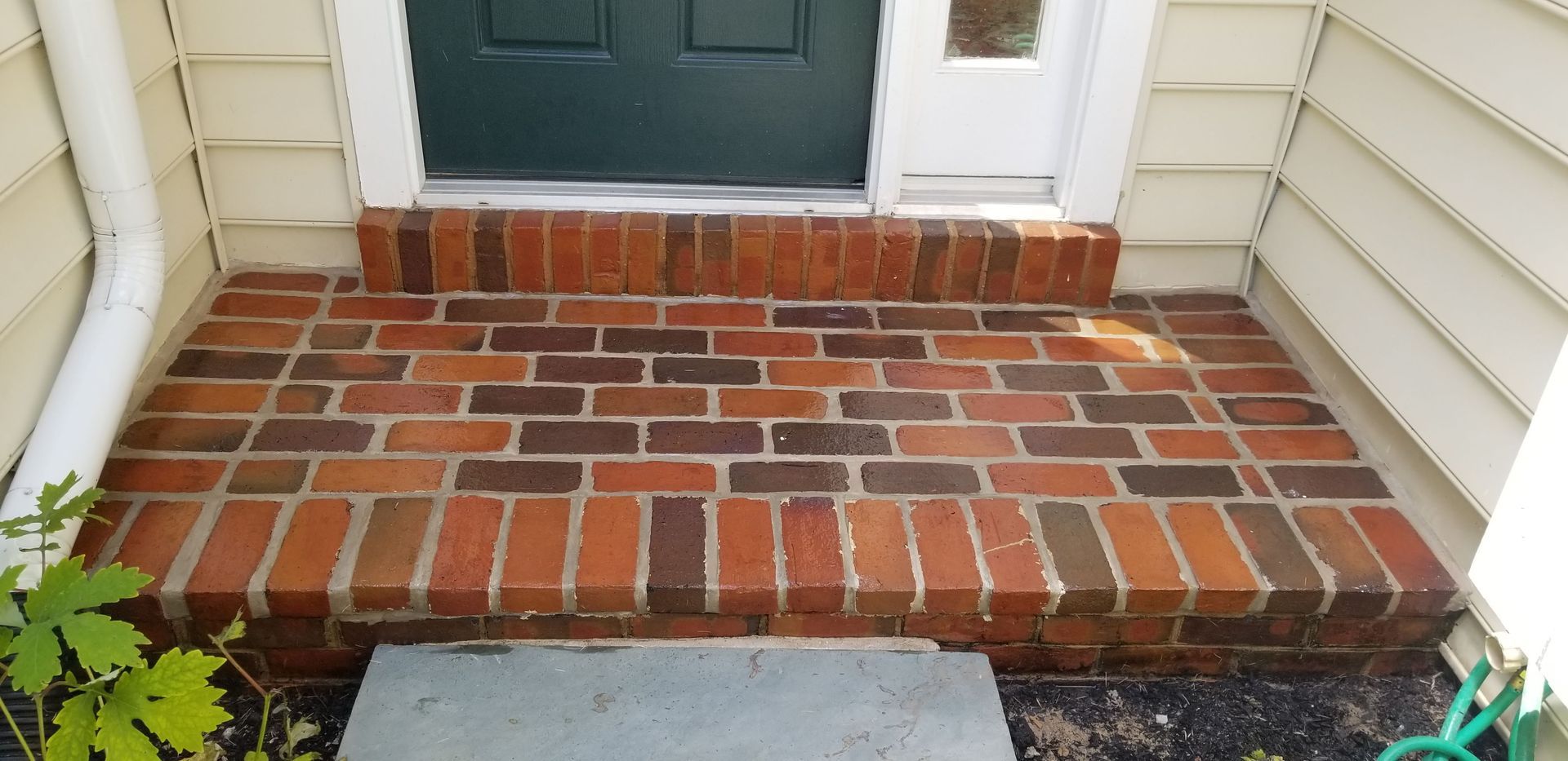 A brick-paved front porch leading to a green door, with light-colored siding on the walls to either side.