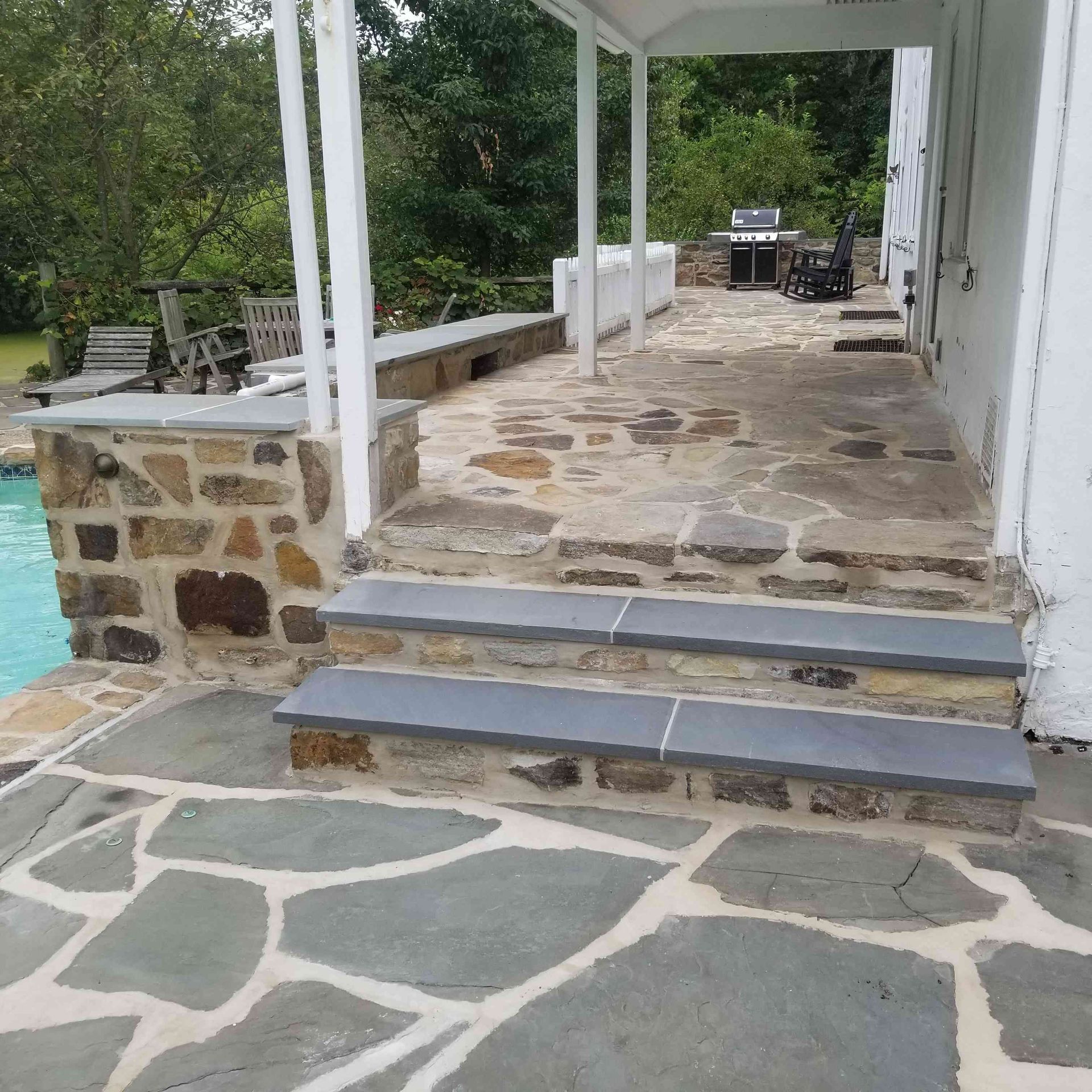 Flagstone patio with two dark stone steps leading to a covered walkway beside a stone wall and swimming pool.