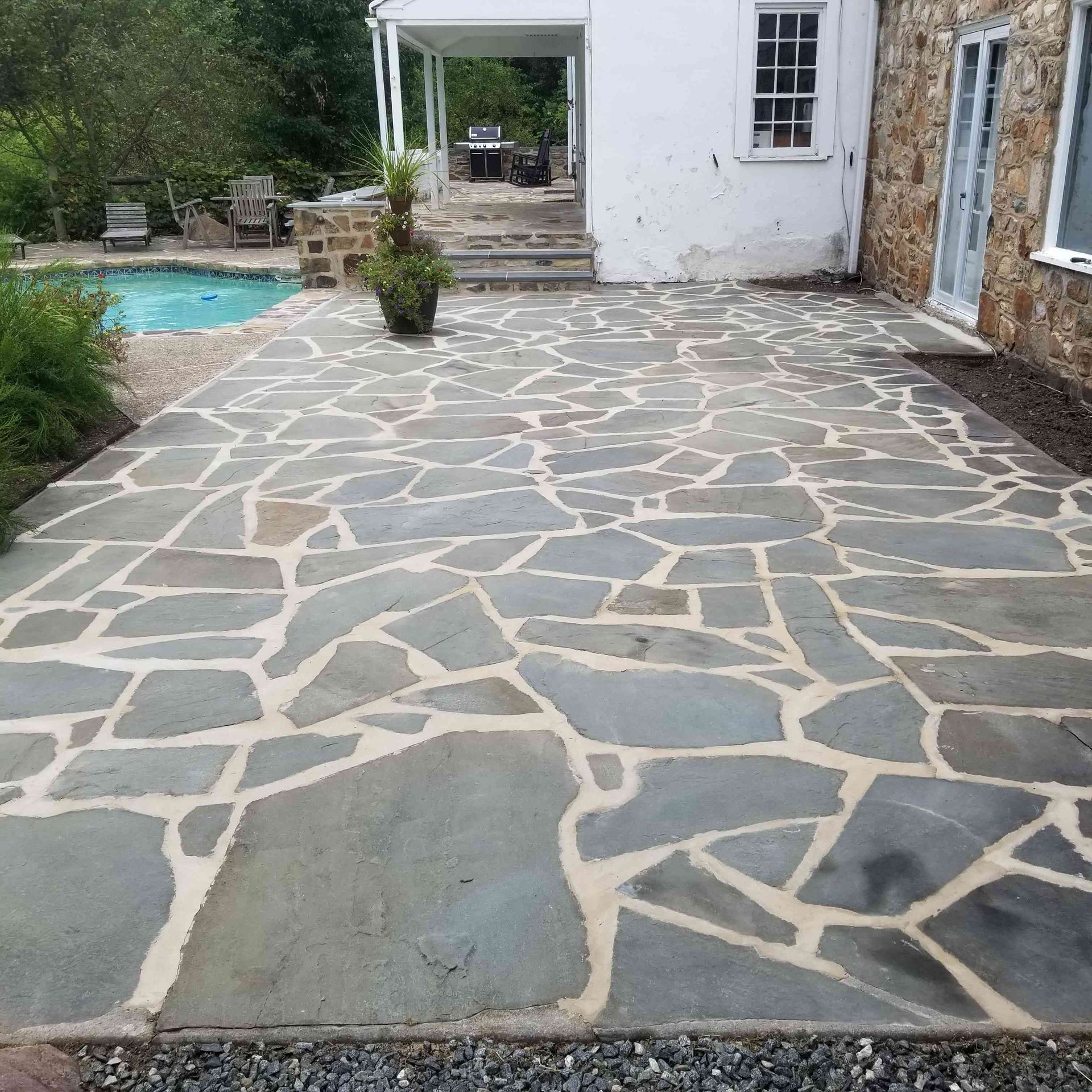 A grey slate flagstone patio with light mortar joints leads to a white house with a swimming pool nearby.