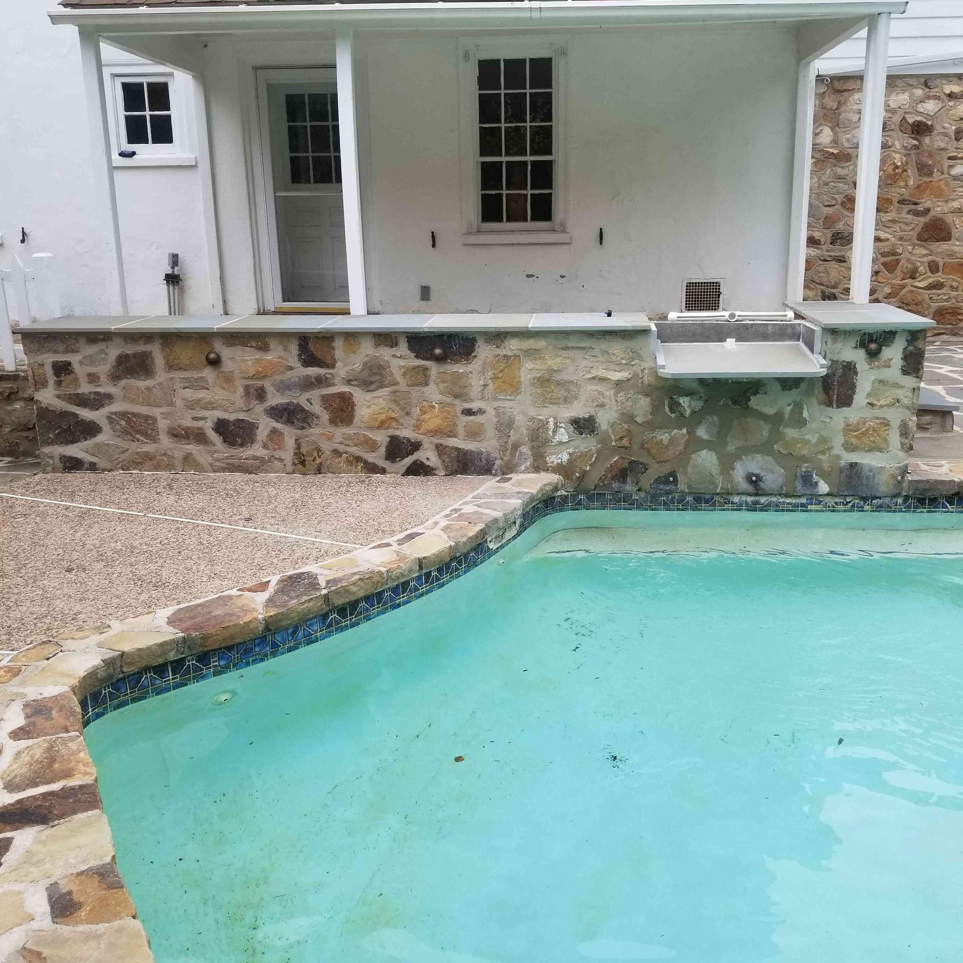 A stone patio wall with a built-in counter next to a backyard swimming pool by a white house.