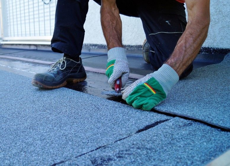 Person in gloves cutting roofing material on a flat roof, dark colors.