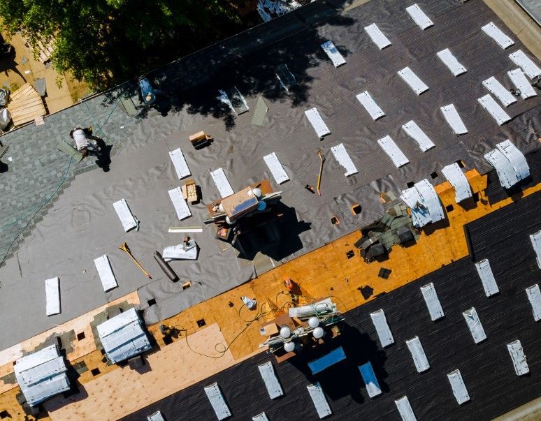 Aerial view of a roof under construction; workers, tools, tar paper and shingles are visible on the roof.