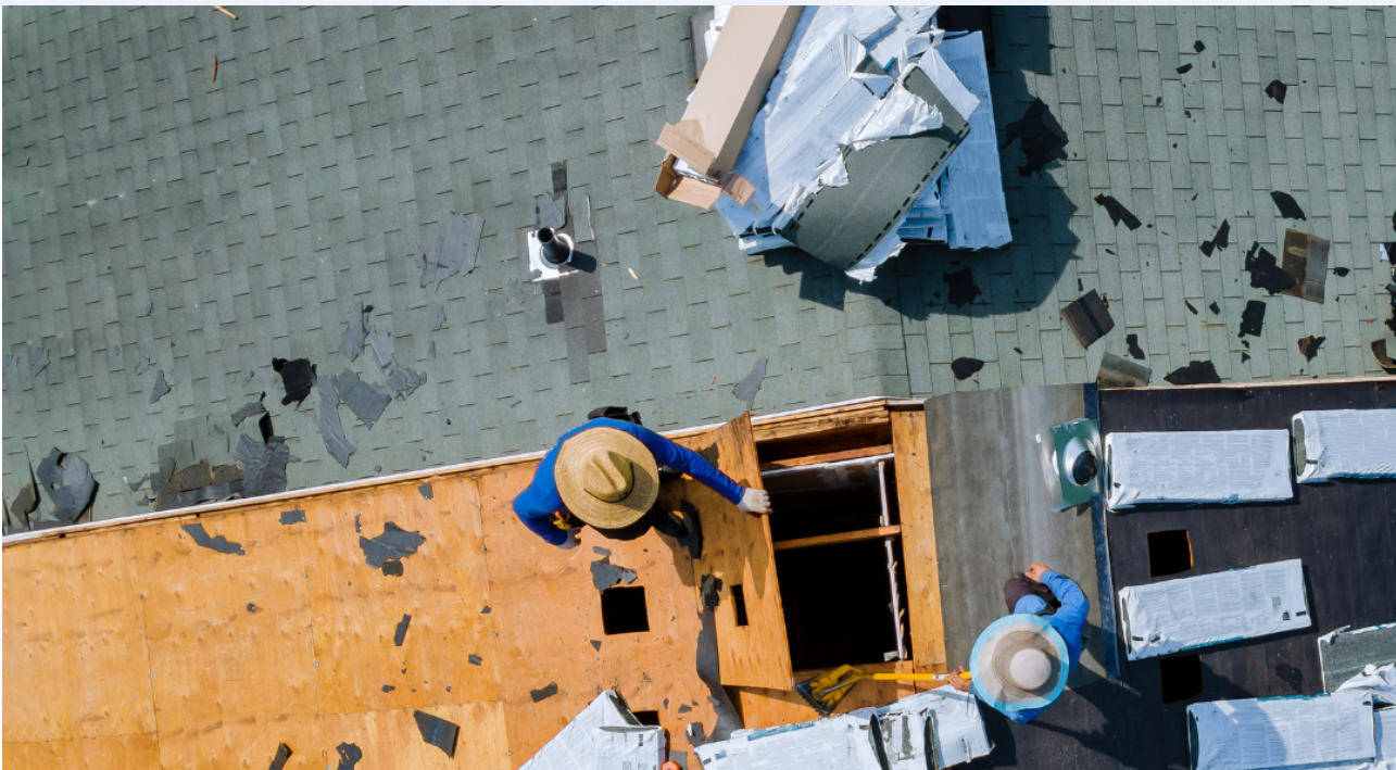 Roofers replacing shingles on a rooftop, viewed from above. One roofer is removing old shingles while another stands nearby.
